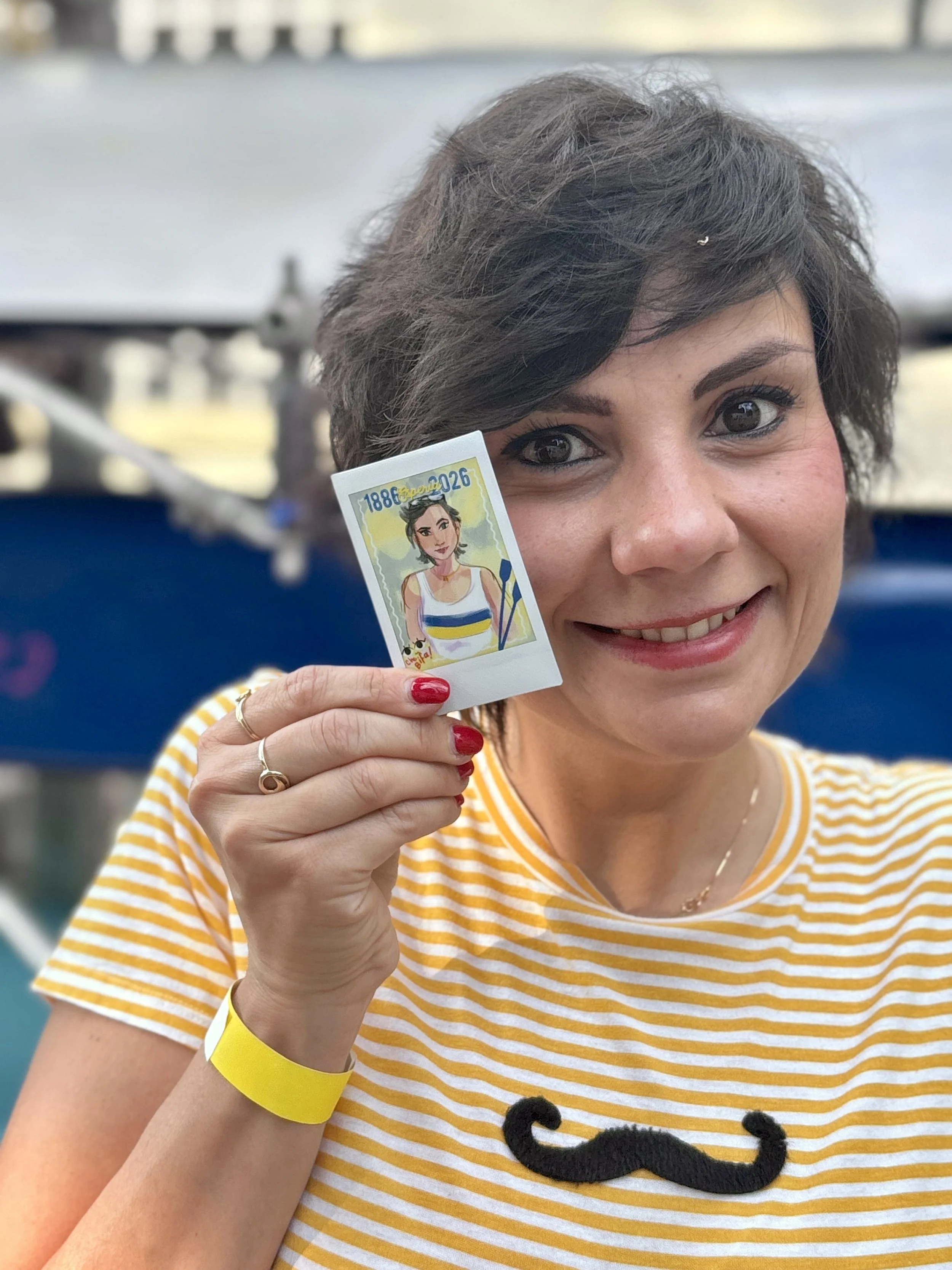 A smiling woman with short curly brown hair holding a ticket with a cartoon illustration of herself. She is wearing a yellow and white striped shirt with a black mustache design, red nail polish, and a yellow wristband.