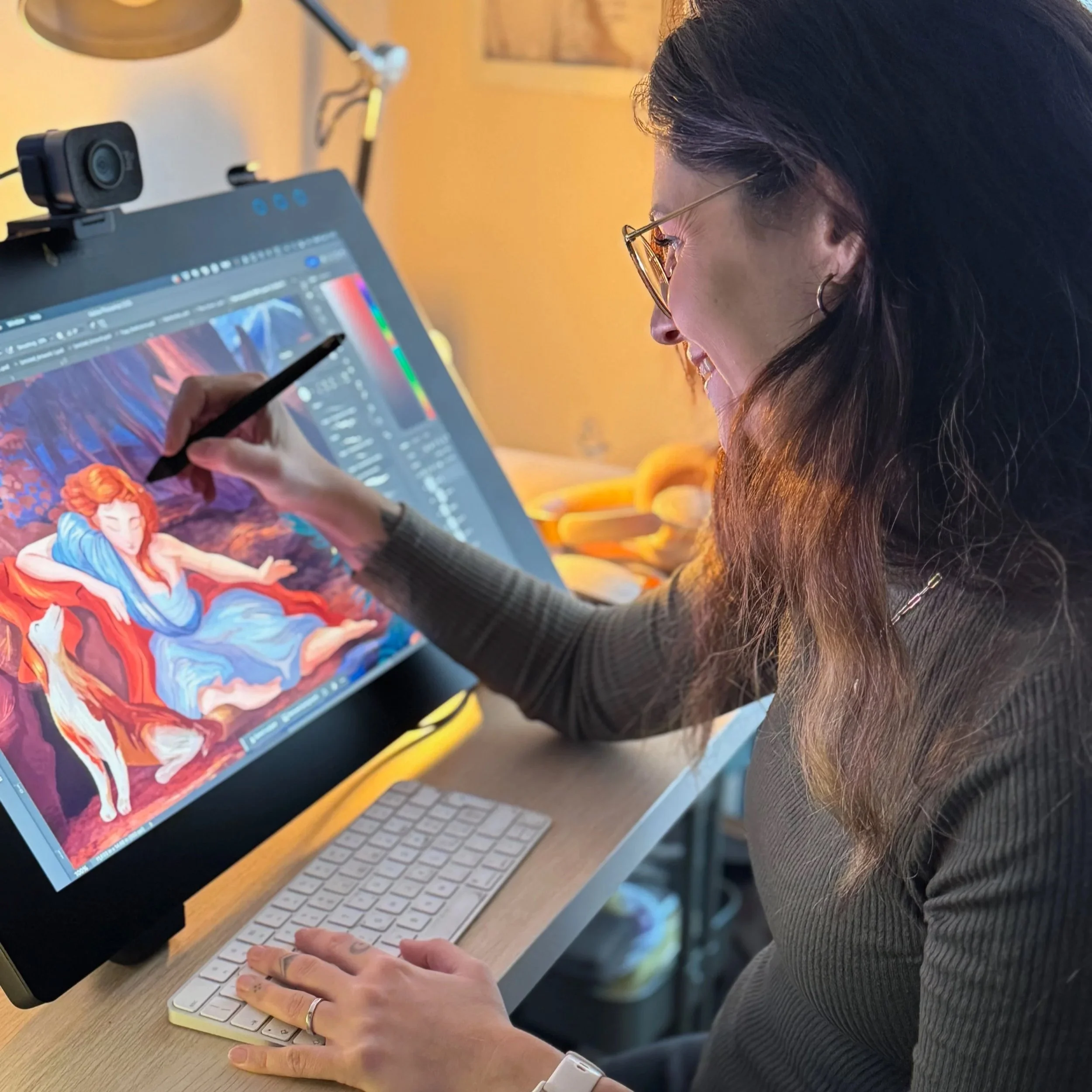 A person wearing glasses and a gray sweater working on a digital art project on a computer screen, which displays colorful animated characters and editing software. They are holding a stylus pen and using a keyboard at a wooden desk, with a camera mounted on the monitor and a yellow cup in the background.
