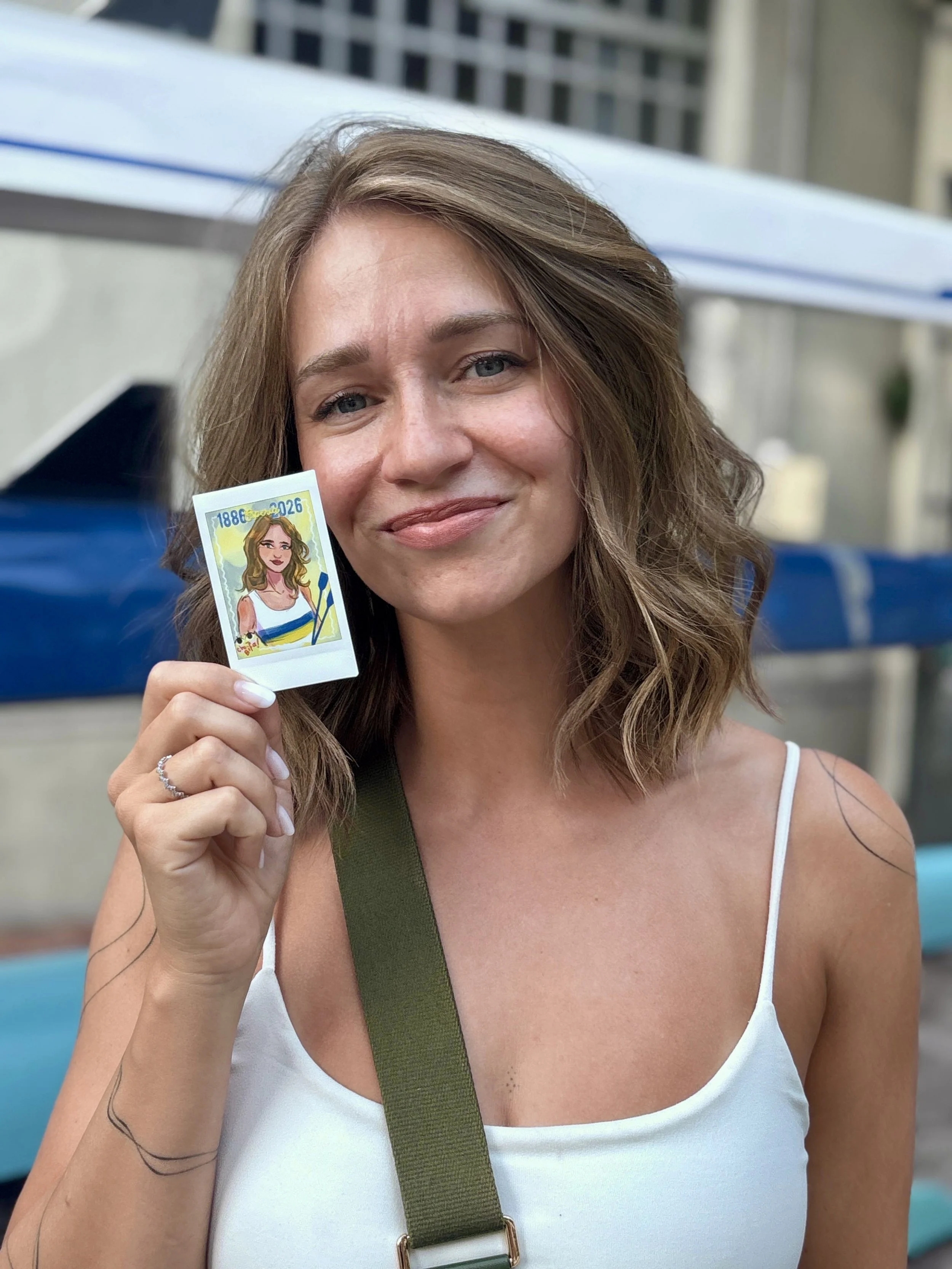 A woman with wavy brown hair and blue eyes is smiling and holding a colorful illustrated sticker depicting a cartoon woman with long hair, wearing a striped top. She is outdoors near a building, wearing a white spaghetti strap top and a green strap b
