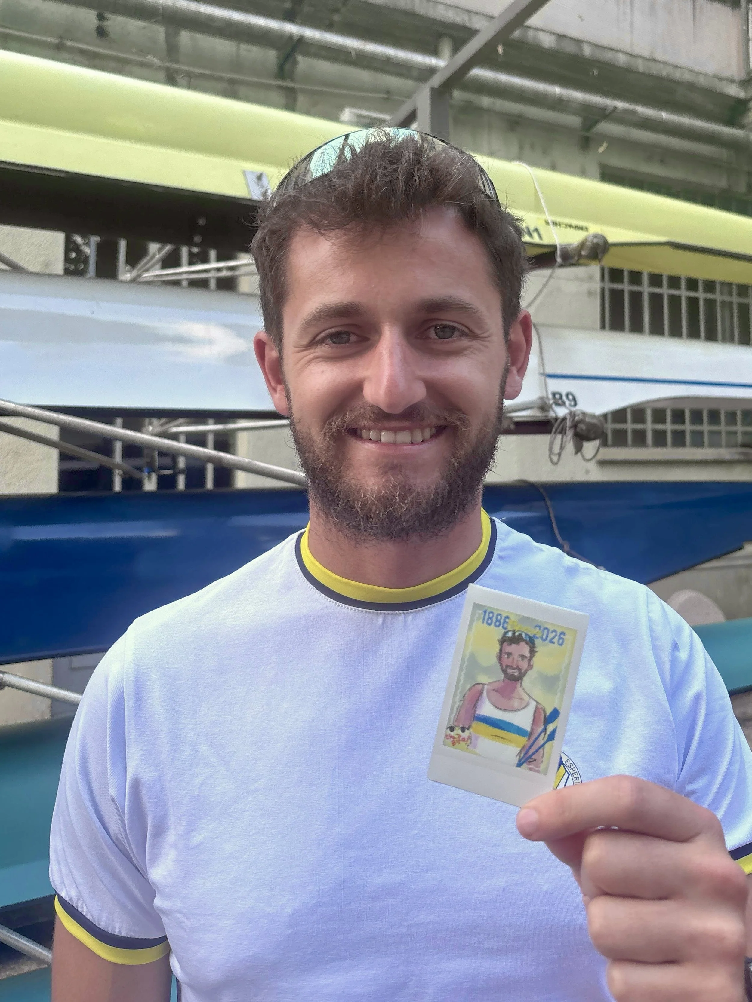 A man holding a paper souvenir card with a cartoon drawing of himself in a tank top, smiling. The man is smiling, wearing a white shirt with yellow trim, and standing in front of a backdrop with boats or kayaks.