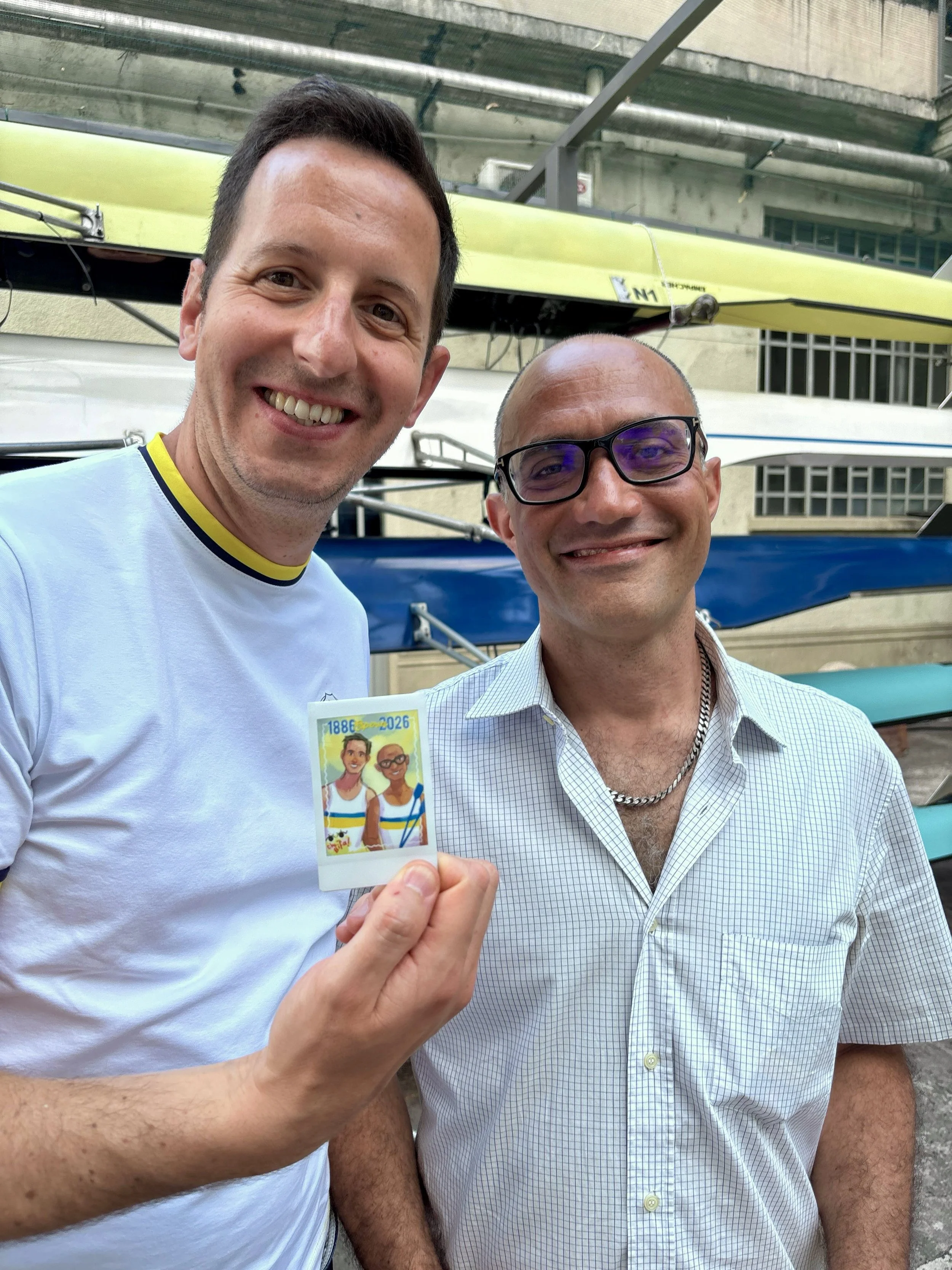 Two men smiling for a selfie outdoors, one holding a colorful sticker with cartoon characters. There are kayaks stacked in the background.