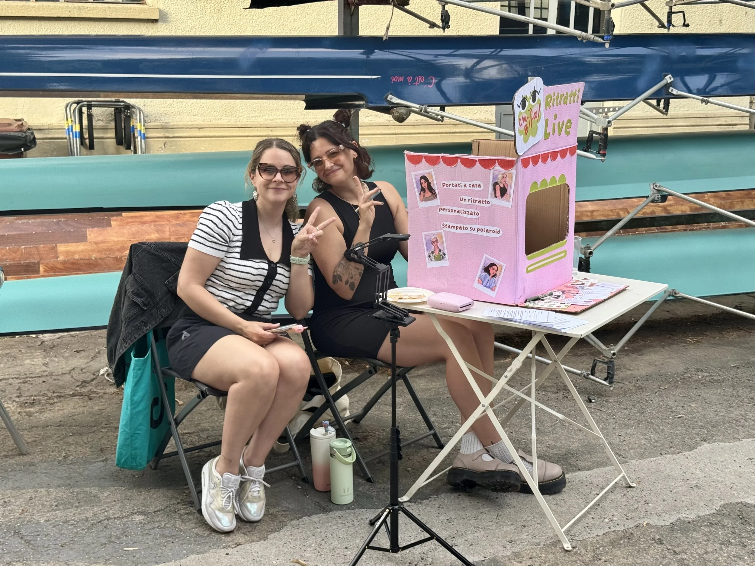 Two women sitting at a table in front of a pink booth, making peace signs and smiling. The booth has signage with illustrations and text in Italian, and there are various items on the table, including papers and cups.