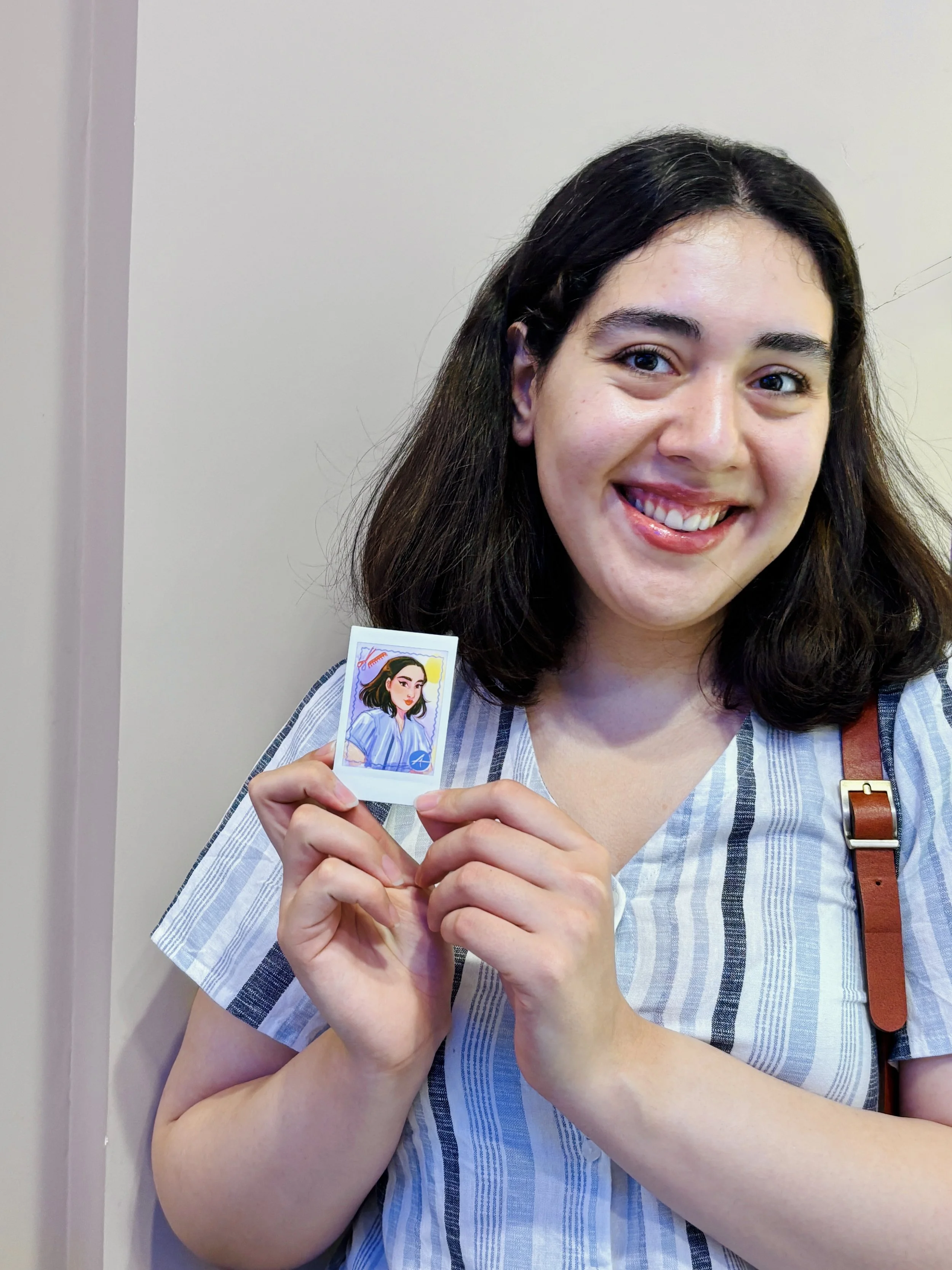 A woman with dark hair, wearing a striped shirt and carrying a brown strap bag, smiling and holding a souvenir photo of herself.