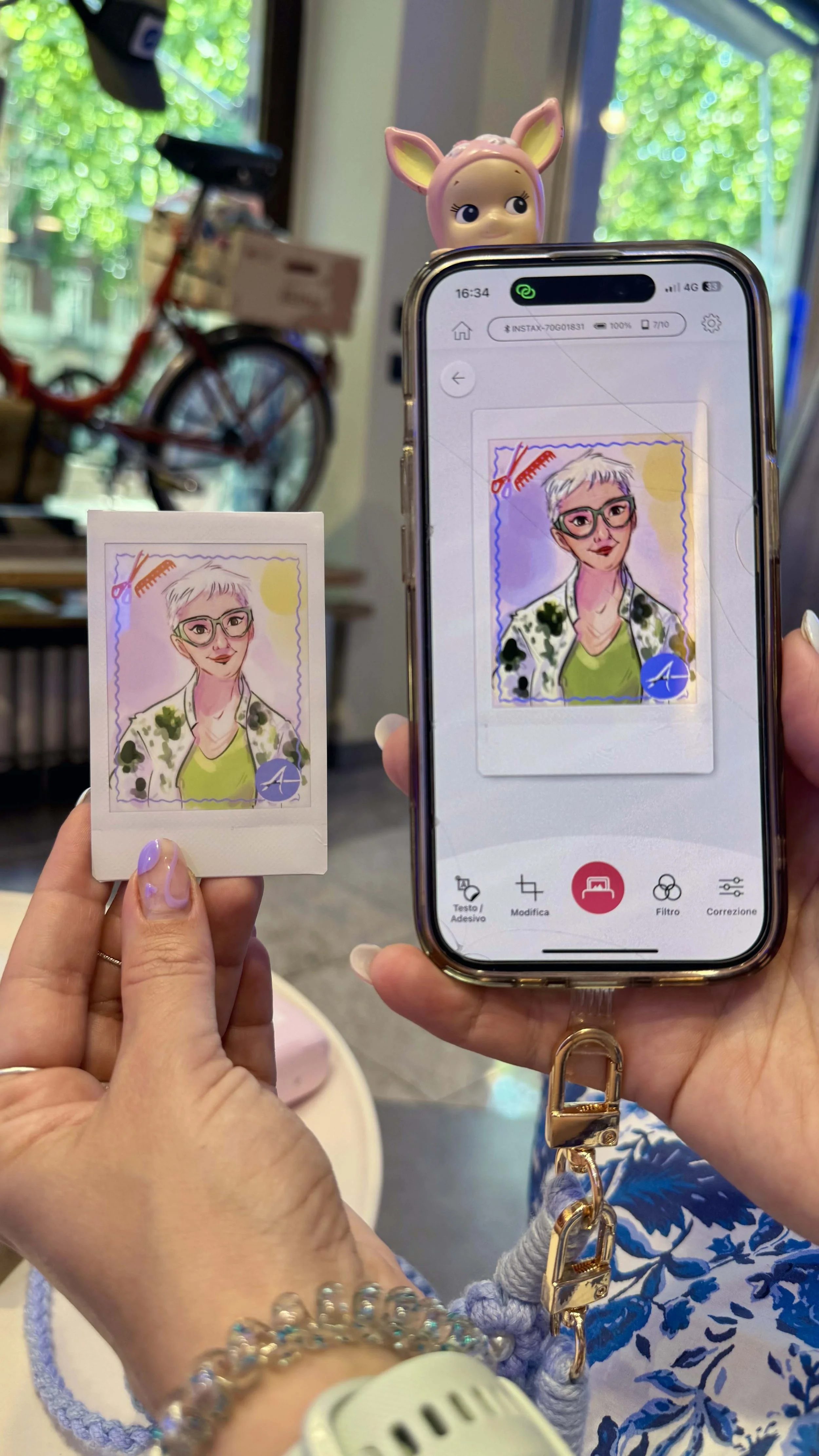 A person is holding a printed cartoon-style portrait of a woman with short white hair, glasses, and a green top, in front of a smartphone displaying a similar digitally edited version of the portrait. The person's hand has a bracelet made of colorful beads, and they are wearing a light-colored watch. In the background, there is a toy with a deer face on top of the smartphone, a bicycle, and a window with blurry green trees outside.