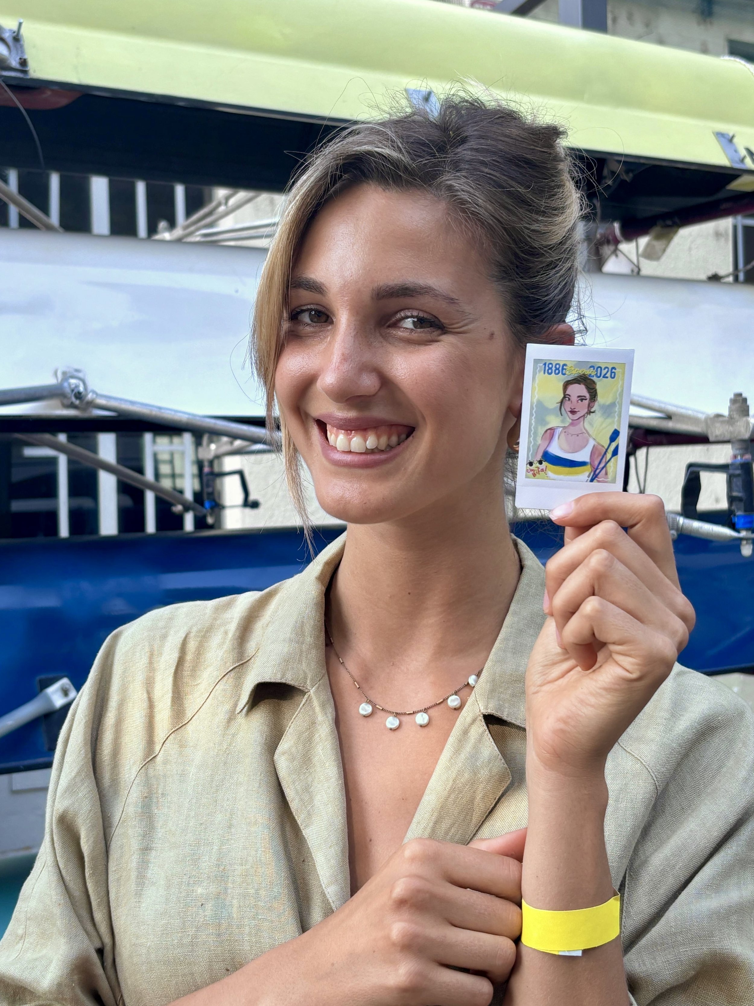 A smiling woman with short brown hair and light makeup, holding a small illustration of a girl with a yellow and blue top and the years 1886-2026. She wears a beige shirt, a pearl necklace, and a yellow wristband, standing outdoors with metal structu