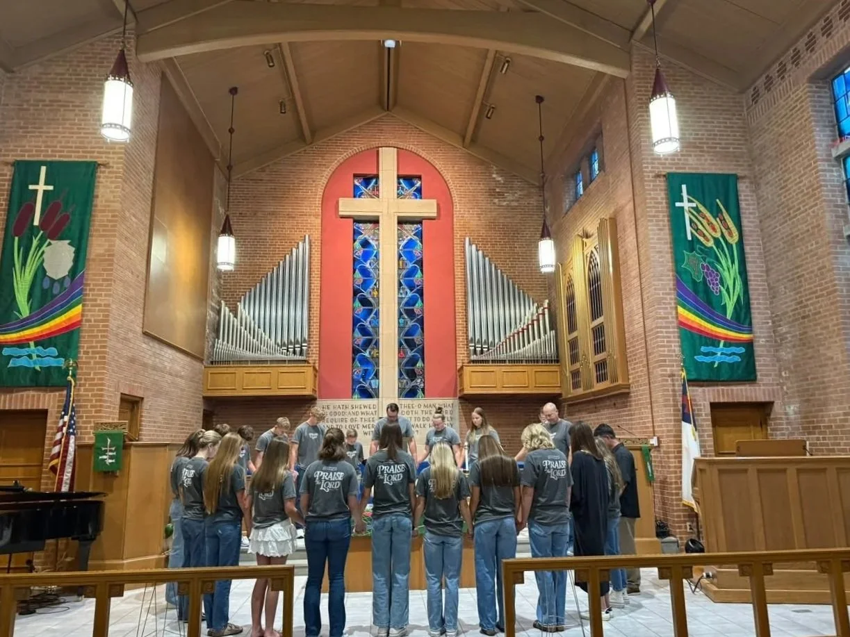 Group of people gathered inside a church, standing in a circle during a prayer or reflection, with a large cross and pipe organ in the background.