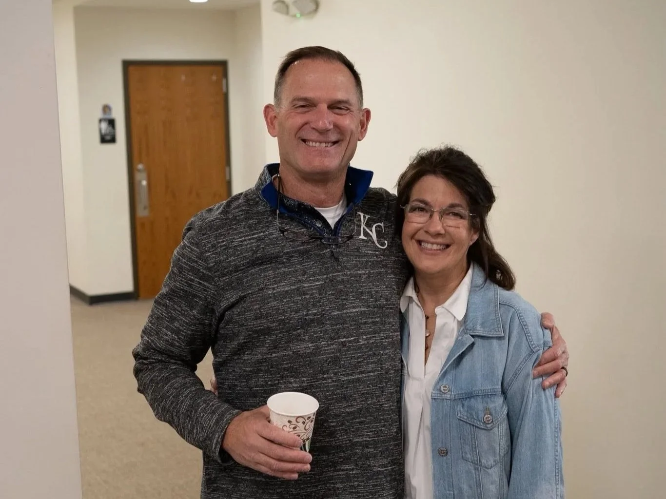 A man and woman standing together indoors, smiling. The man is holding a cup. The man is wearing a dark gray pullover with a white LS with a small logo. The woman is wearing glasses, a white shirt, and a light blue denim jacket.