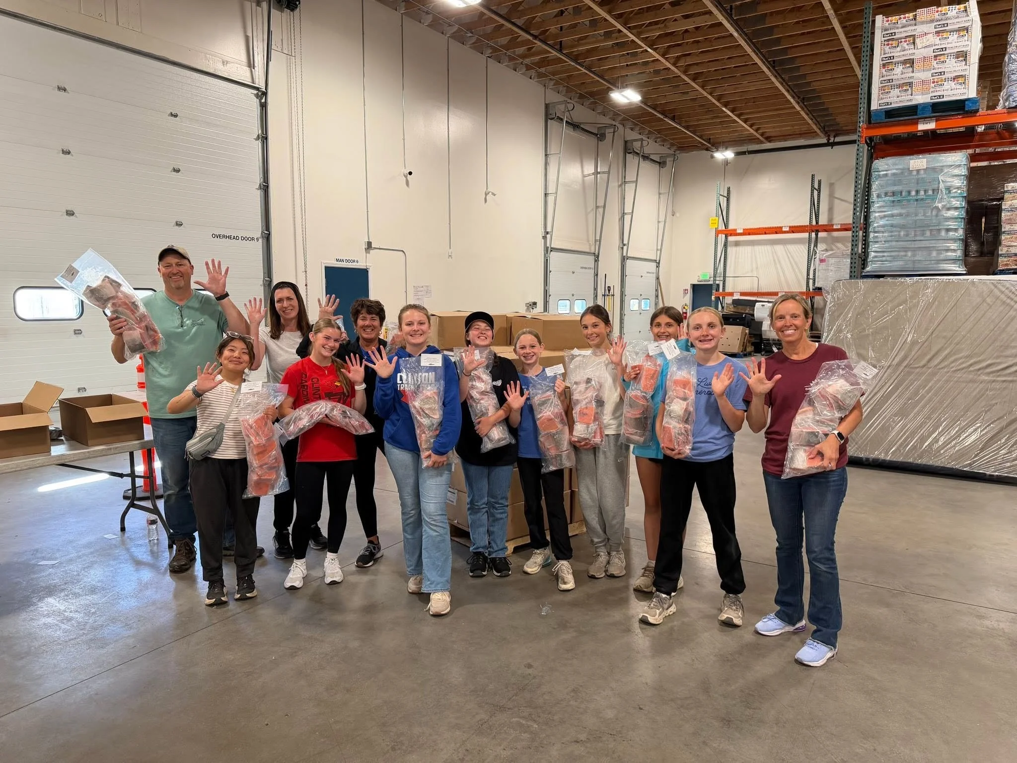 Group of people, mostly young girls, posing with their hands raised inside a warehouse, holding packages, with shelves and boxes in the background.