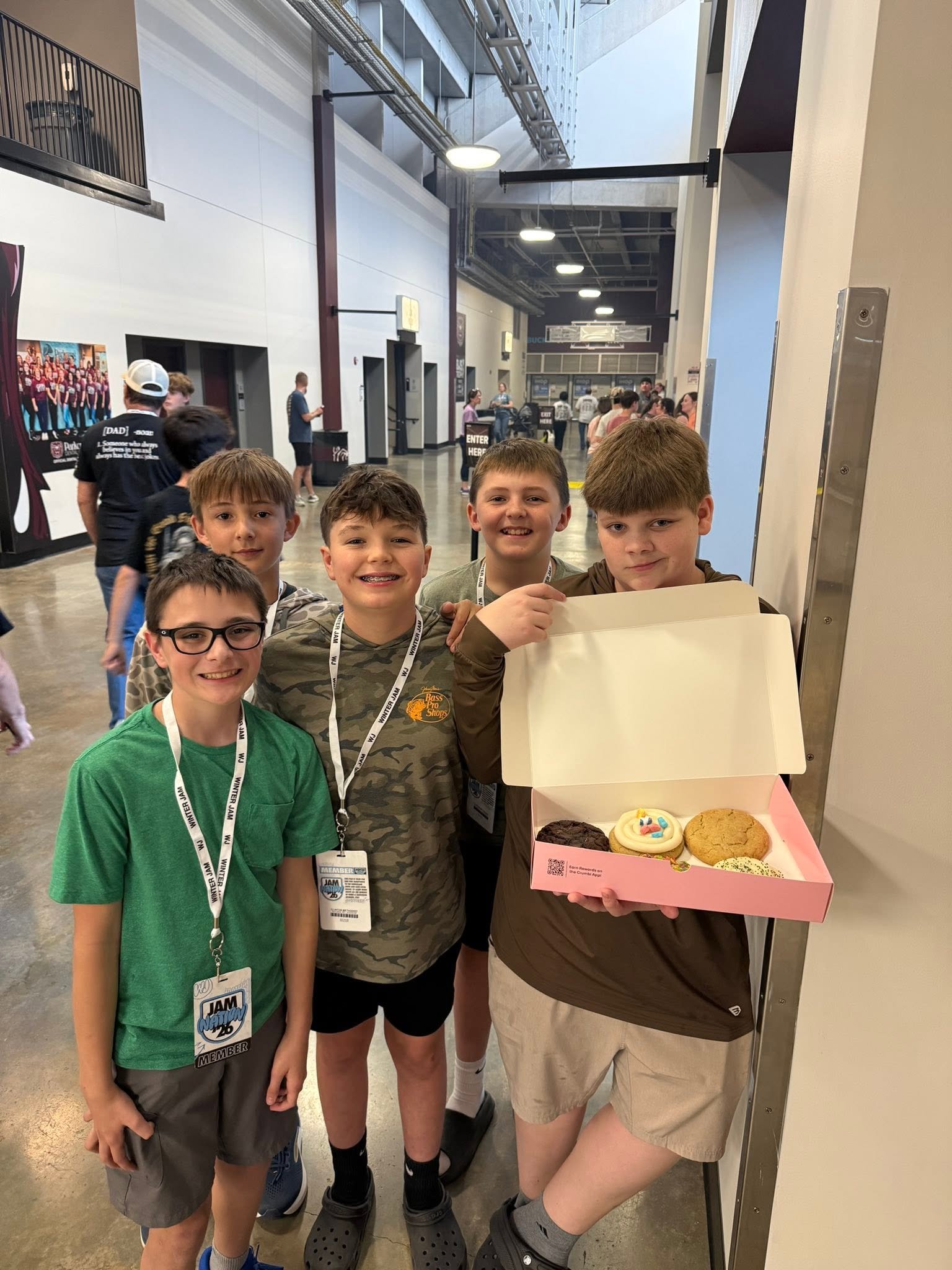 Group of young boys at an indoor event, one holding a box of assorted cookies, standing in line or gathering near a table with other children and adults in the background.