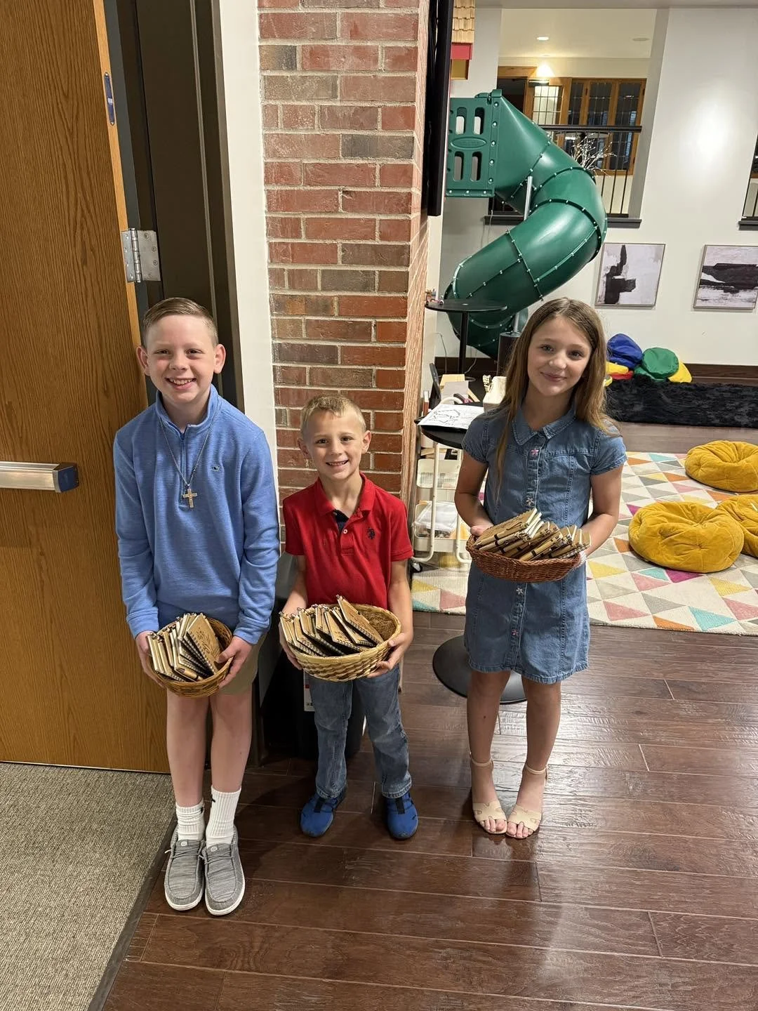 Three children standing inside a building, holding baskets filled with small paper bags. A green spiral slide and colorful cushions are visible in the background.