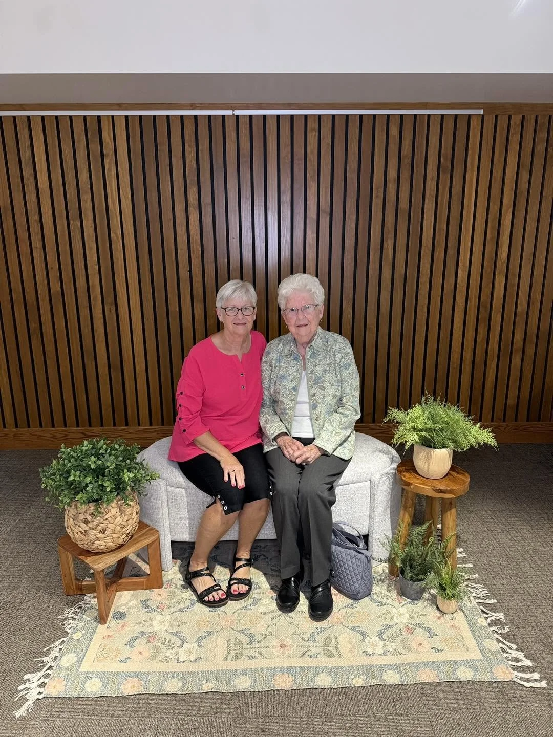 Two elderly women sitting on a light-colored bench with green plants on wooden side tables on either side, against a wooden slatted wall background.