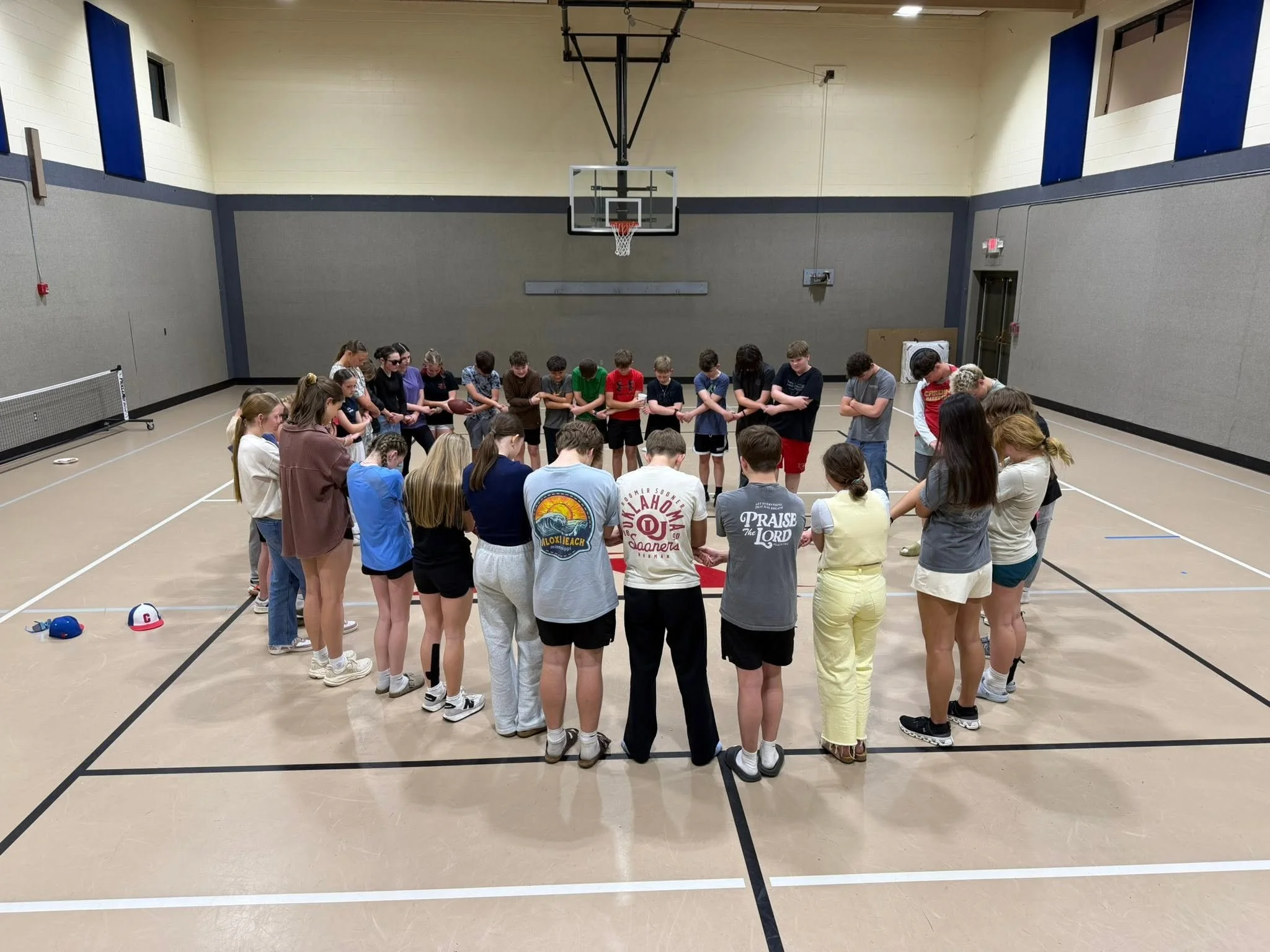 A group of children and teenagers are standing in a circle in a gymnasium, with most of their heads bowed or eyes closed, holding hands or touching shoulders, possibly praying or engaging in a group activity. There is a basketball hoop on the wall be