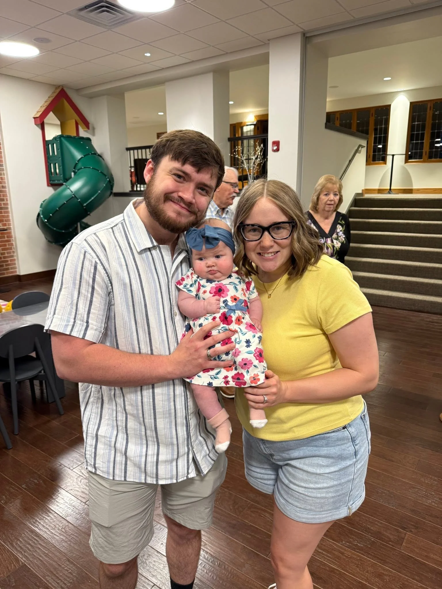 Three adults and one baby girl smiling indoors, with staircase, windows, and a play slide in the background.