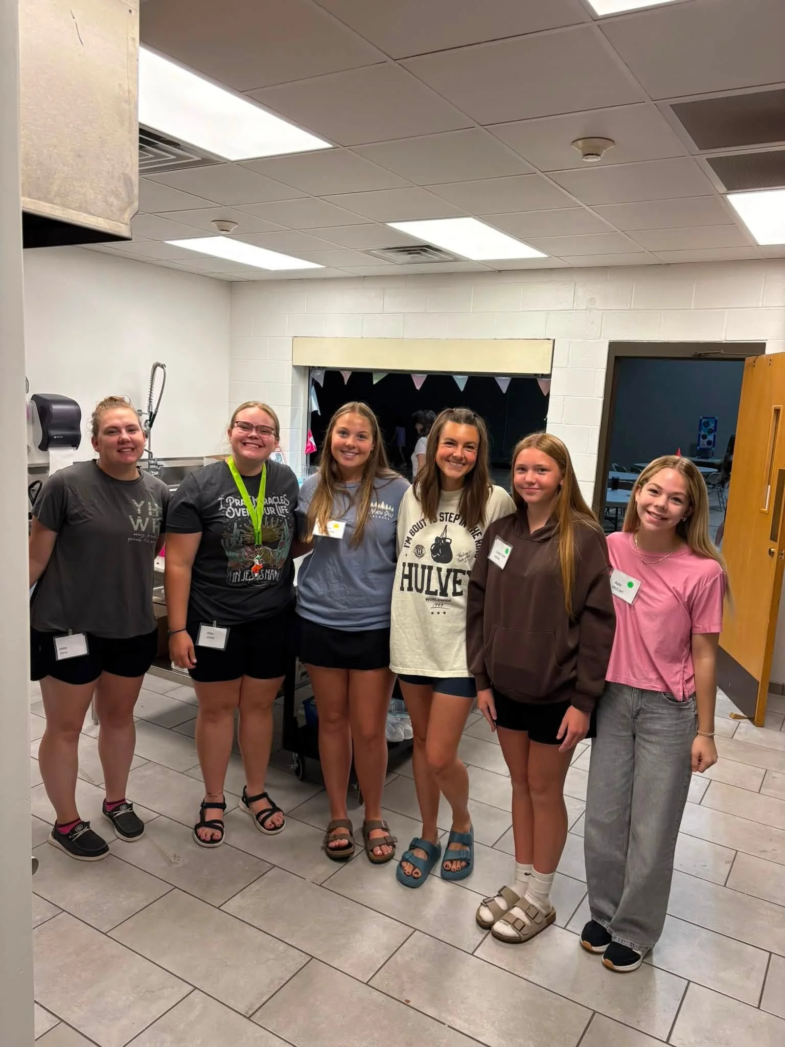 Group of six young girls standing together indoors, smiling at the camera. They are wearing casual clothing and sandals, with name tags. The background includes a window with bunting decoration, a sink, and a door.