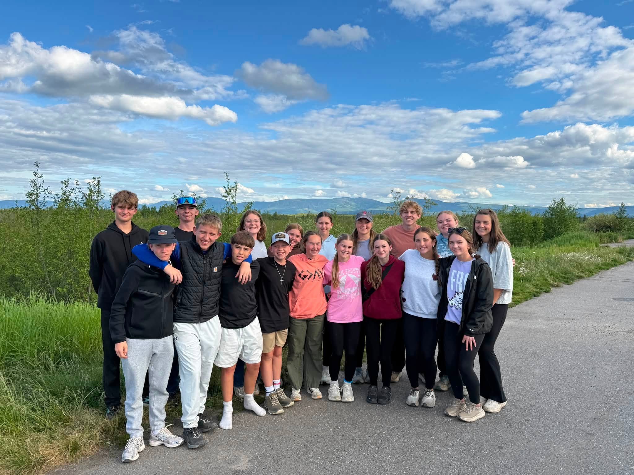 A large group of children and teenagers standing outdoors on a hiking trail with mountains, trees, and a blue sky with clouds in the background.