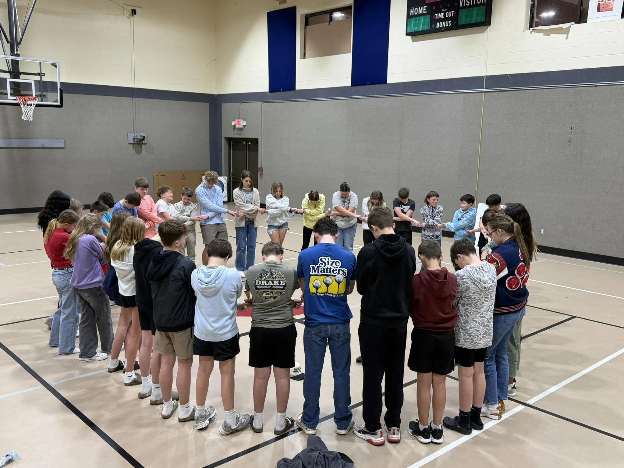A group of children and teenagers standing in a circle holding hands in a gymnasium, participating in an activity or game.