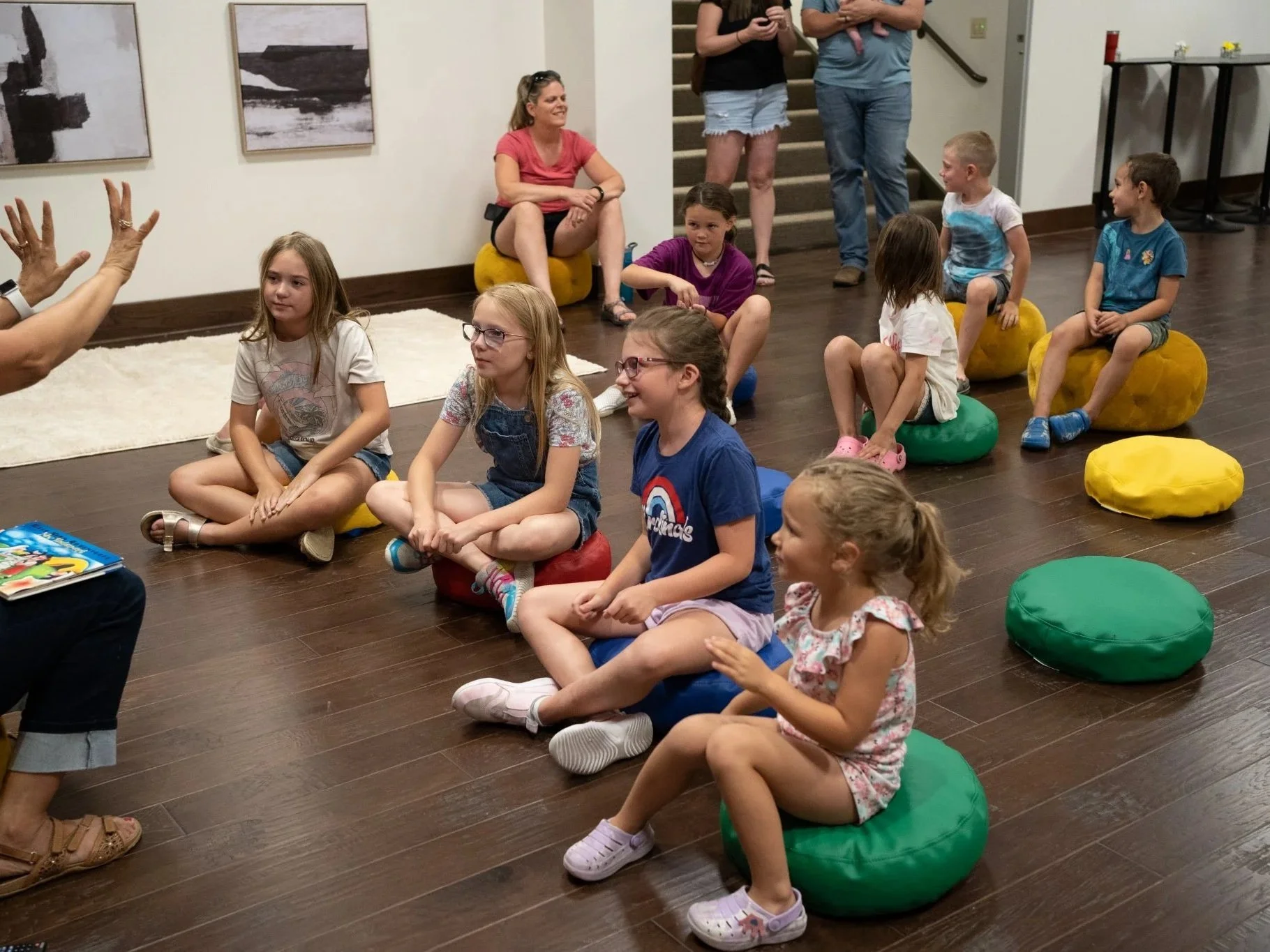 Group of children sitting on colorful round cushions on a wooden floor, participating in an indoor activity, with a woman leading and gesturing with her hands.