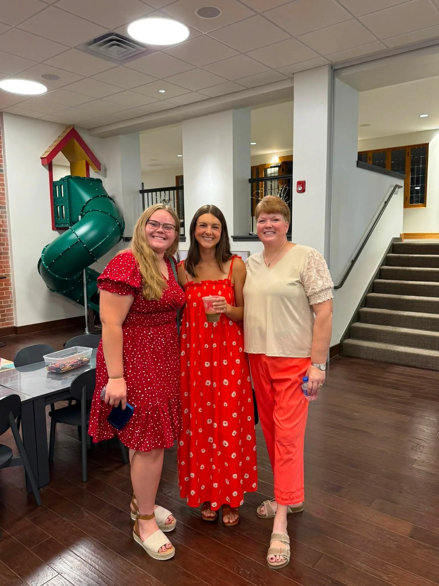Three women standing together indoors, smiling at the camera. Two are wearing red dresses with floral patterns, and one is wearing a cream-colored top with red pants. The women are wearing festive headbands with silver tinsel. There is a green slide outside the window, a staircase to the right, and a table with chairs and a plastic container in the foreground.