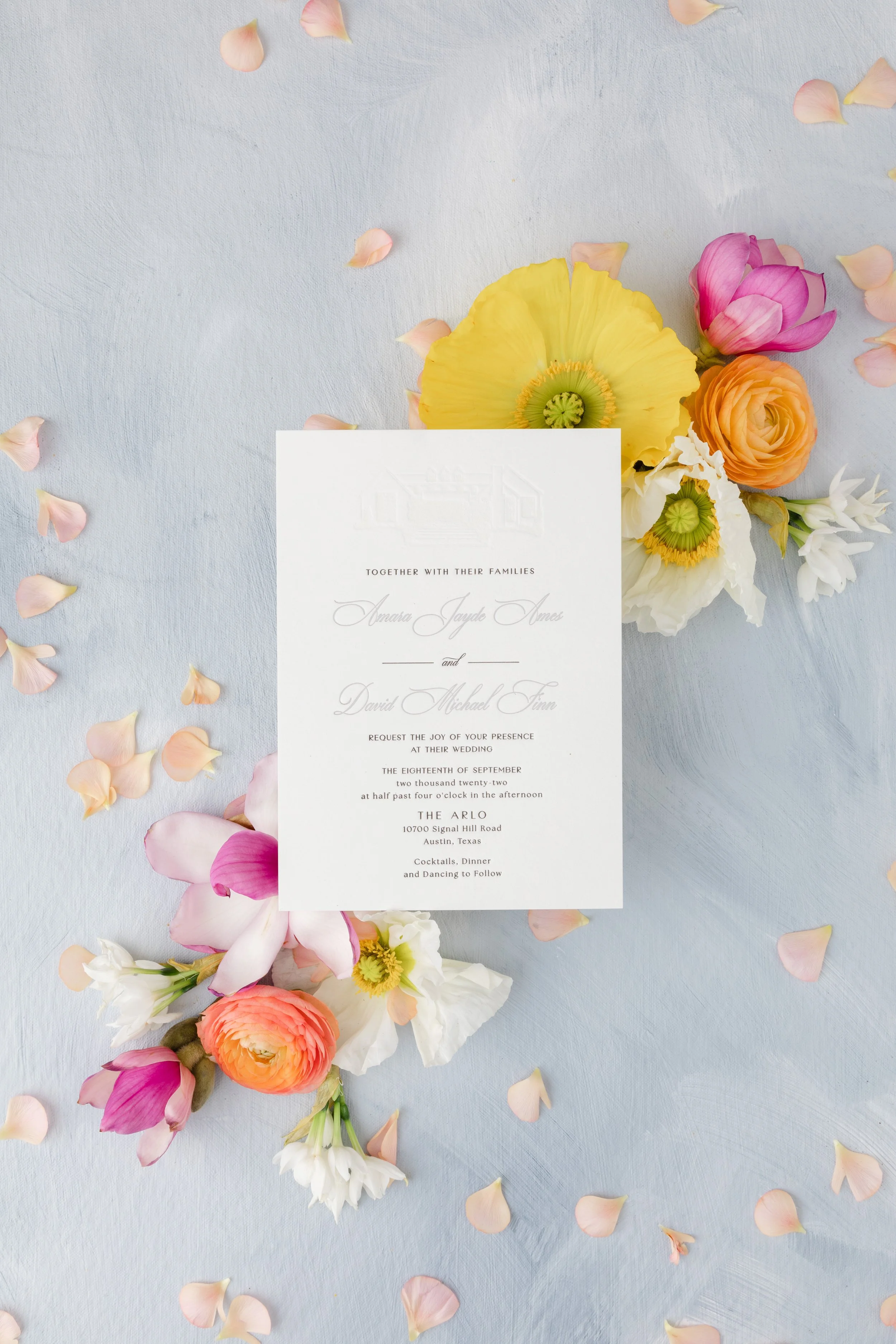 Wedding invitation card on a blue surface surrounded by colorful flowers and scattered pink petals.