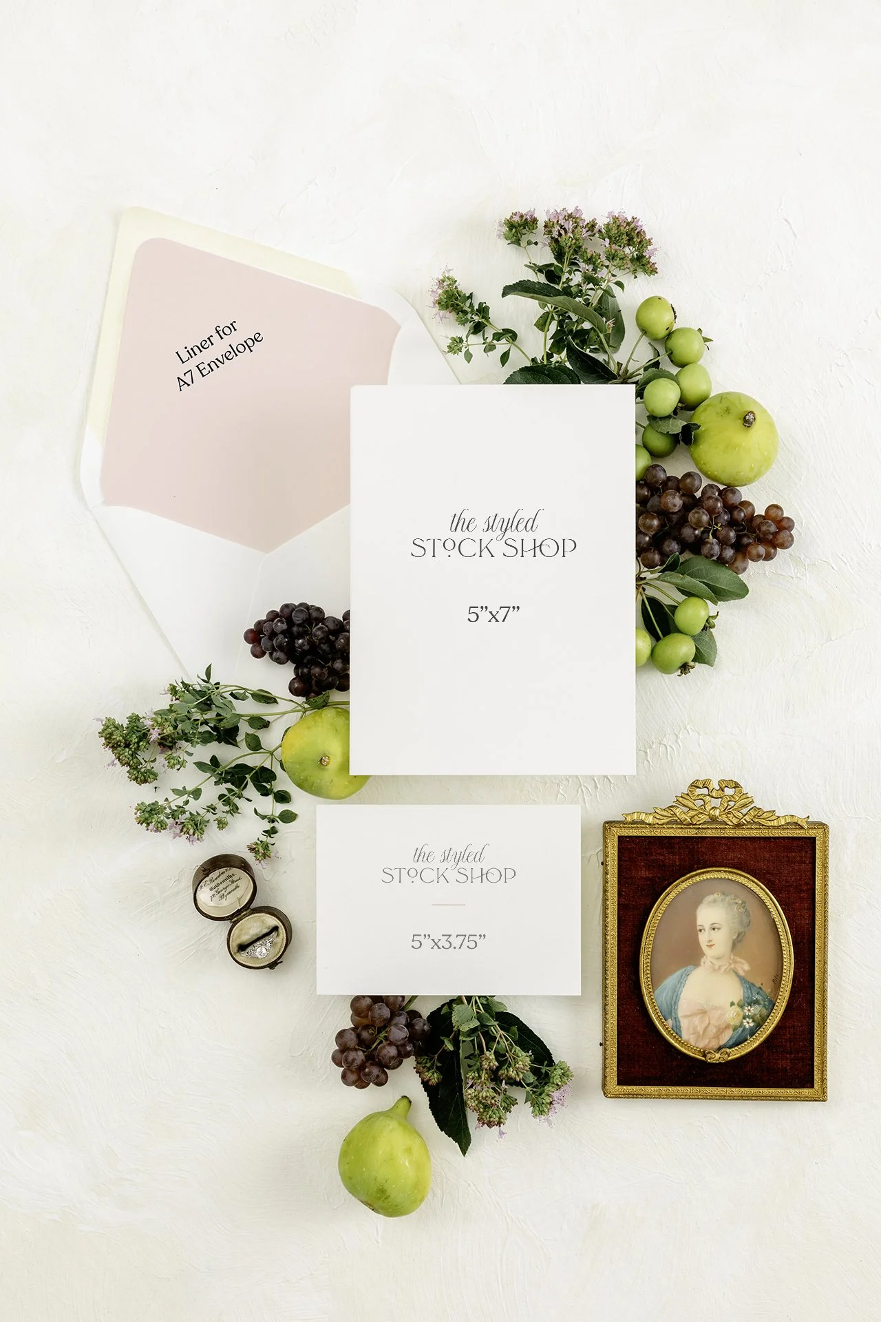 Flat lay of wedding stationery, including a large white card reading 'the styled STOCK SHOP 5'x7"', a smaller card with similar text reading '5"x3.75"', an envelope with a pink liner labeled 'Liner for A7 Envelope', surrounded by grapes, apples, and sprigs of flowers, with a small vintage portrait of a woman in a gilded frame.
