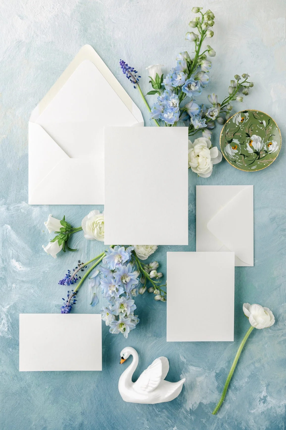 Flat lay of white cards, envelopes, flowers, a small plate, and a ceramic swan on a textured blue surface.
