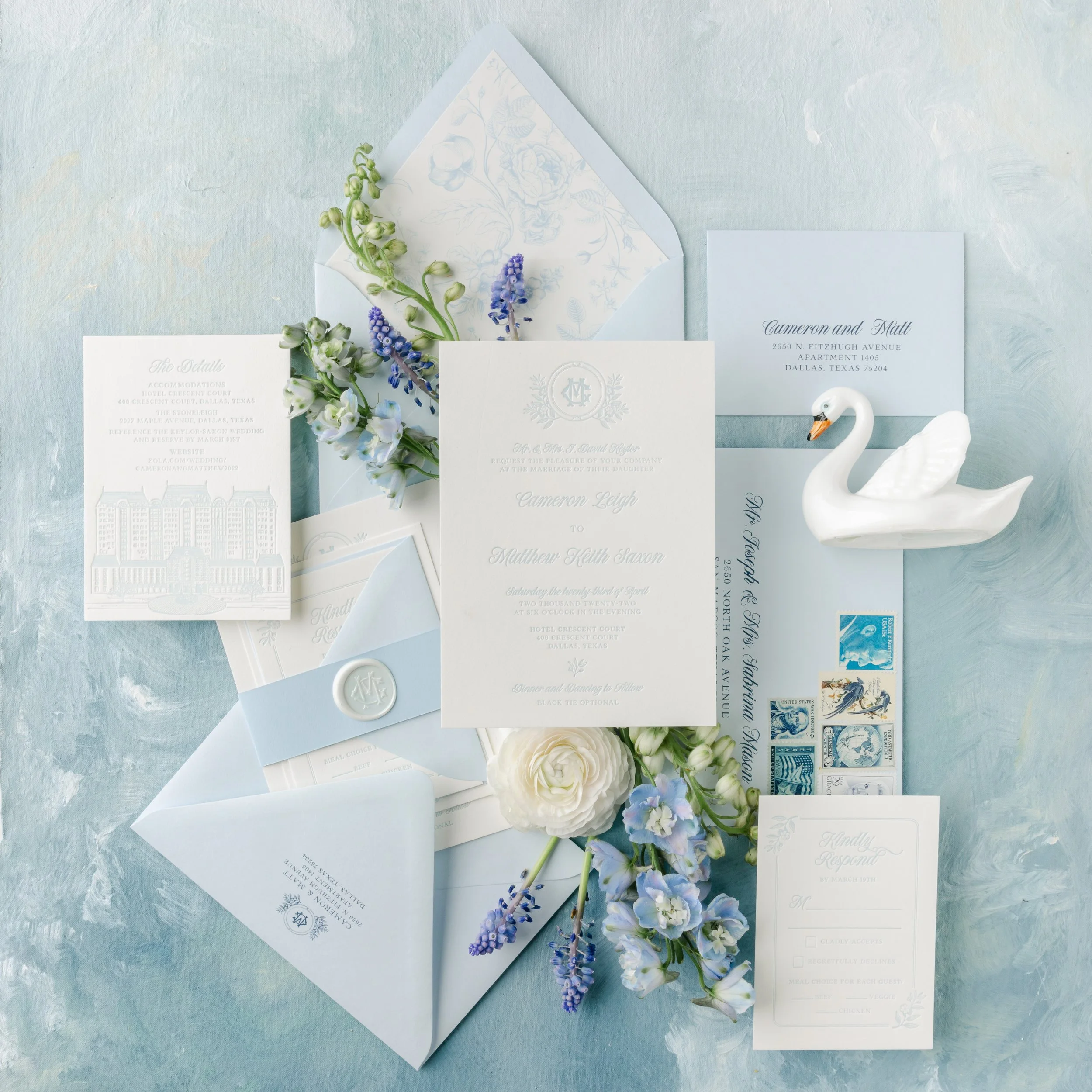 Elegant wedding invitation suite with floral accents, light blue envelopes, and vintage stamps. Includes RSVP card, details card, and a decorative wax seal. White flowers and a ceramic swan are also present.