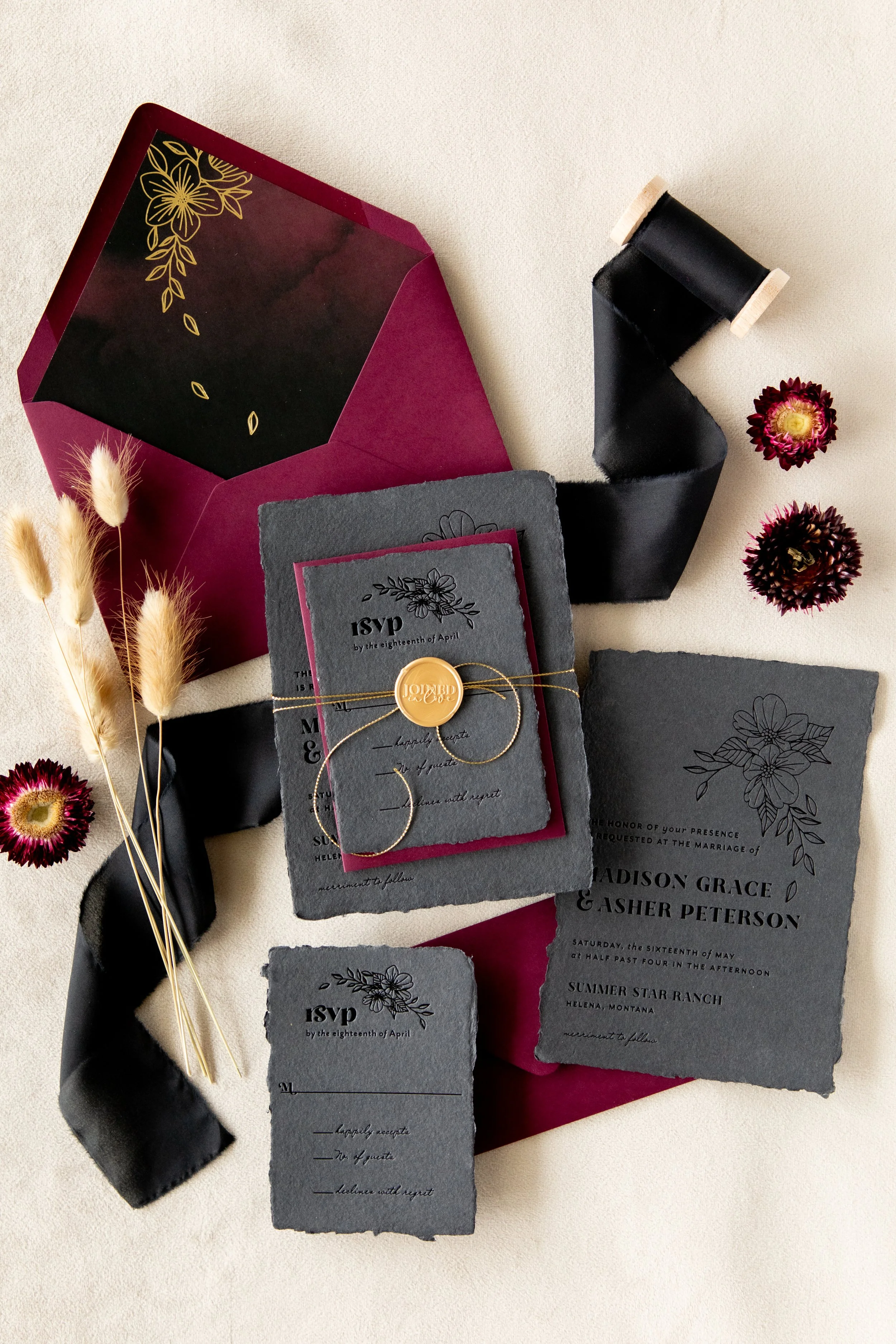Elegant wedding invitation set with dark paper, floral motifs, wax seal, burgundy envelope, black ribbon, dried flowers, and decorative elements.