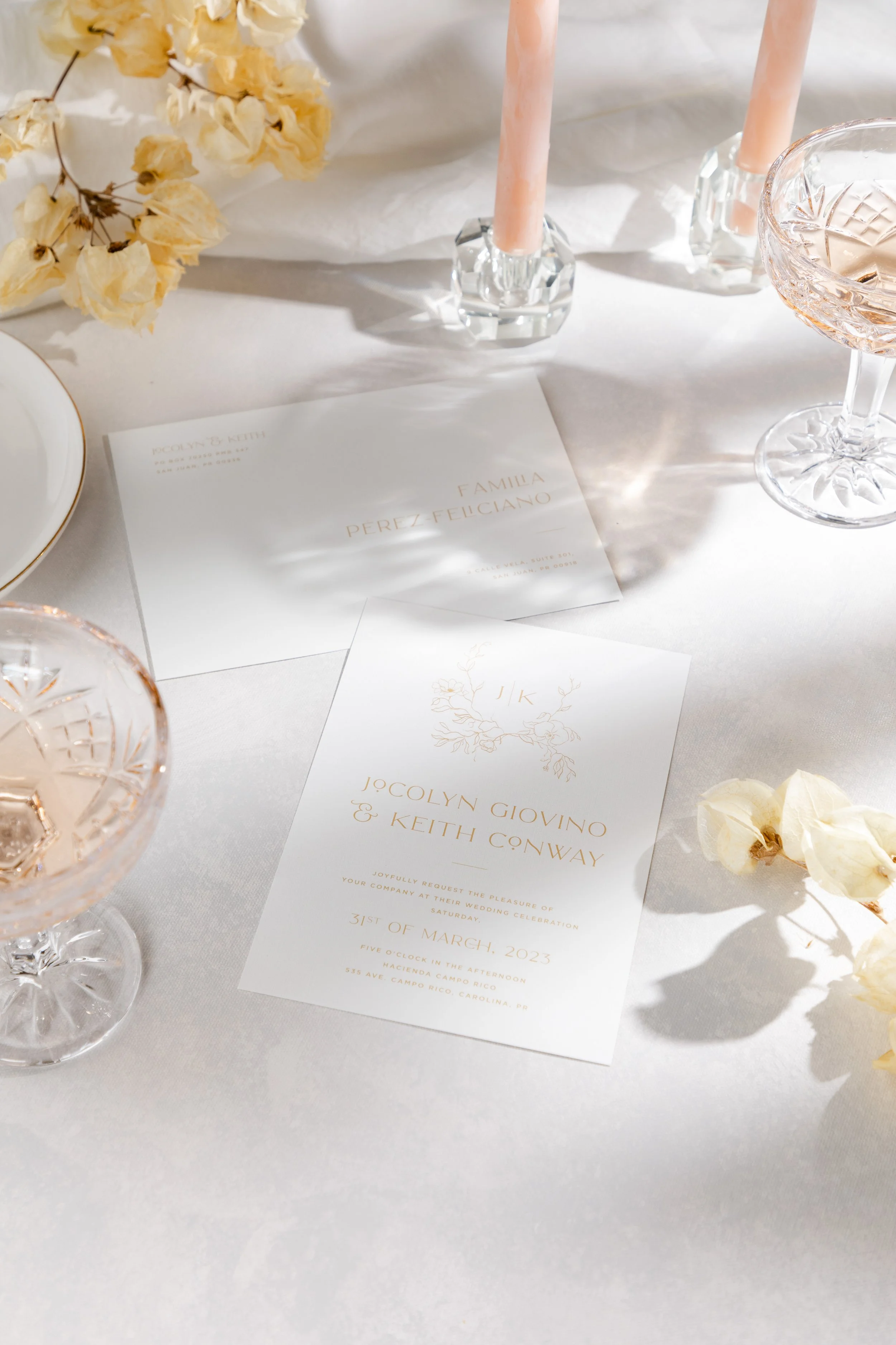 Wedding invitation set on a table with pink candles, dried flowers, and crystal glassware.