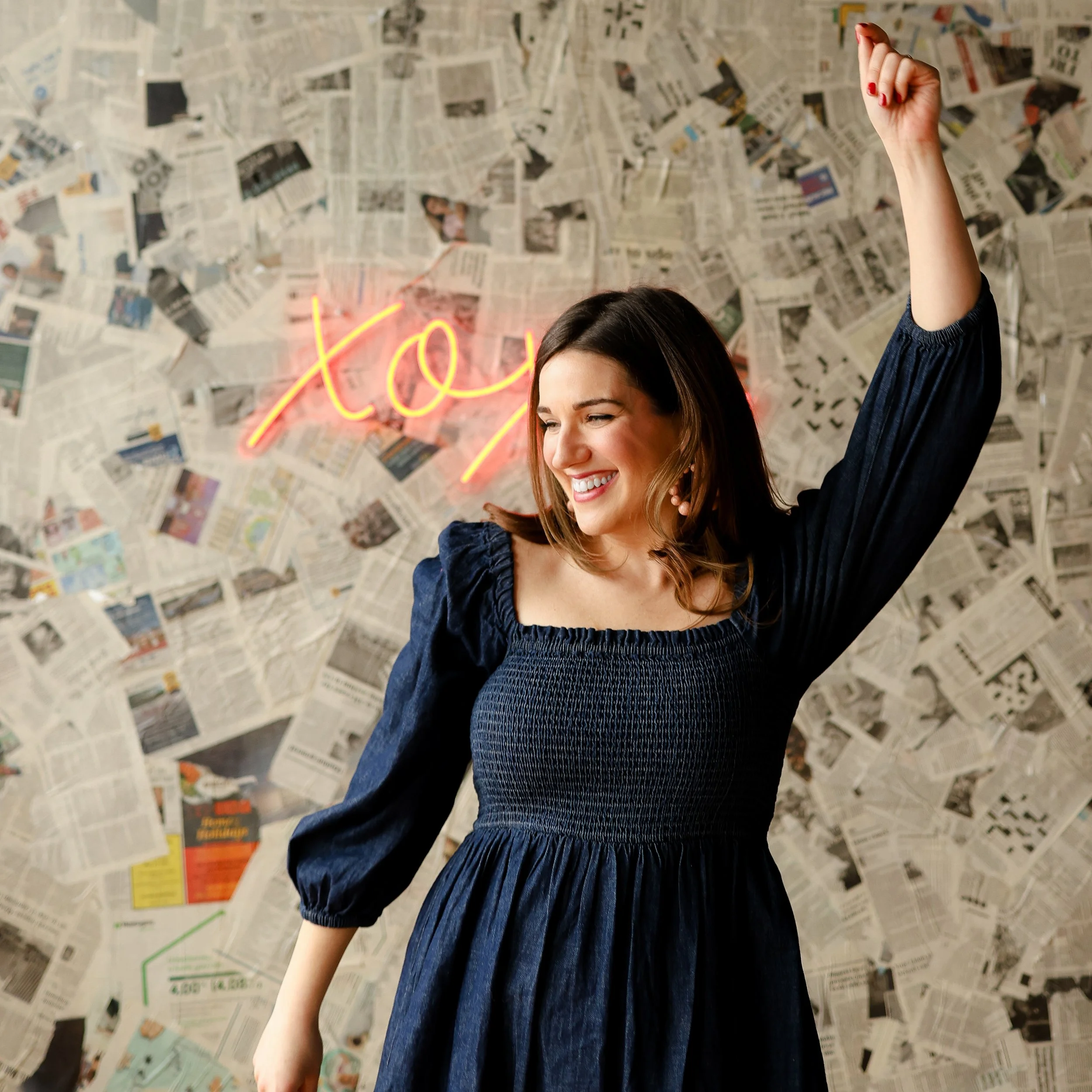 Woman in a dark dress smiling with one arm raised, in front of a wall covered in newspaper clippings; neon XO sign in the background.