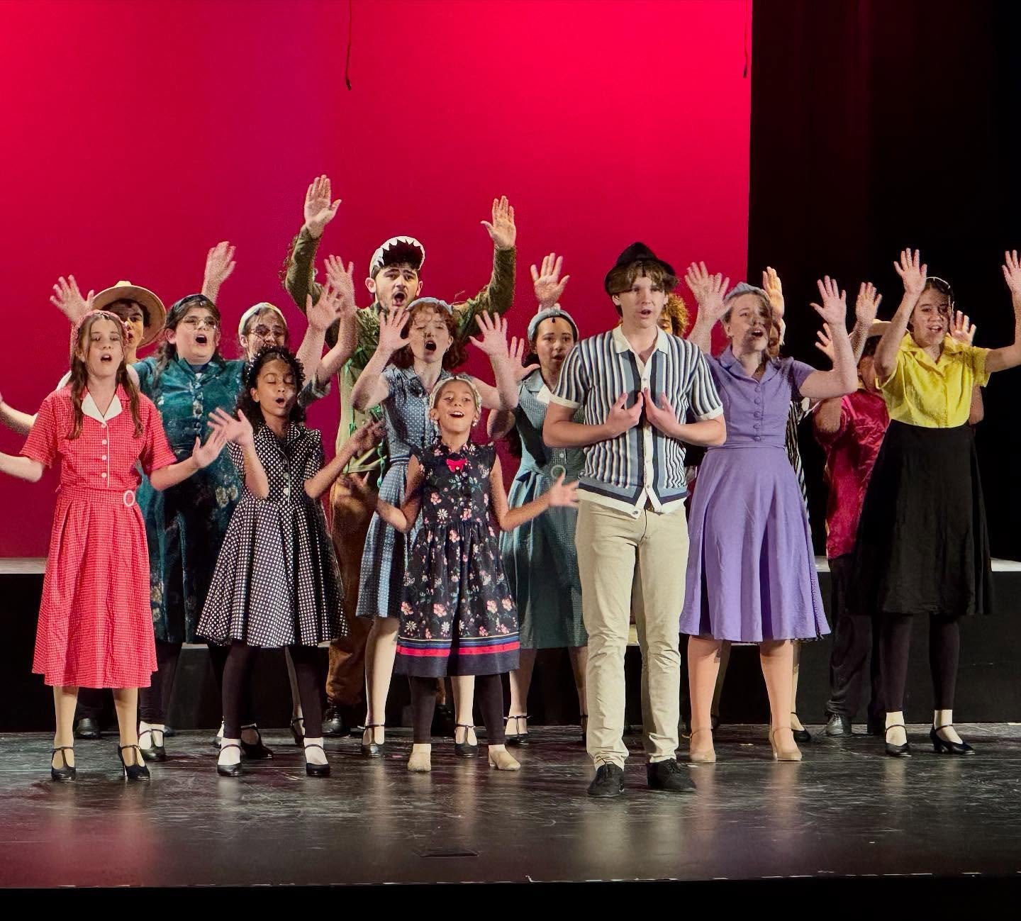 A group of children performing on stage, singing with their hands raised, dressed in colorful retro-style clothing, against a red background.