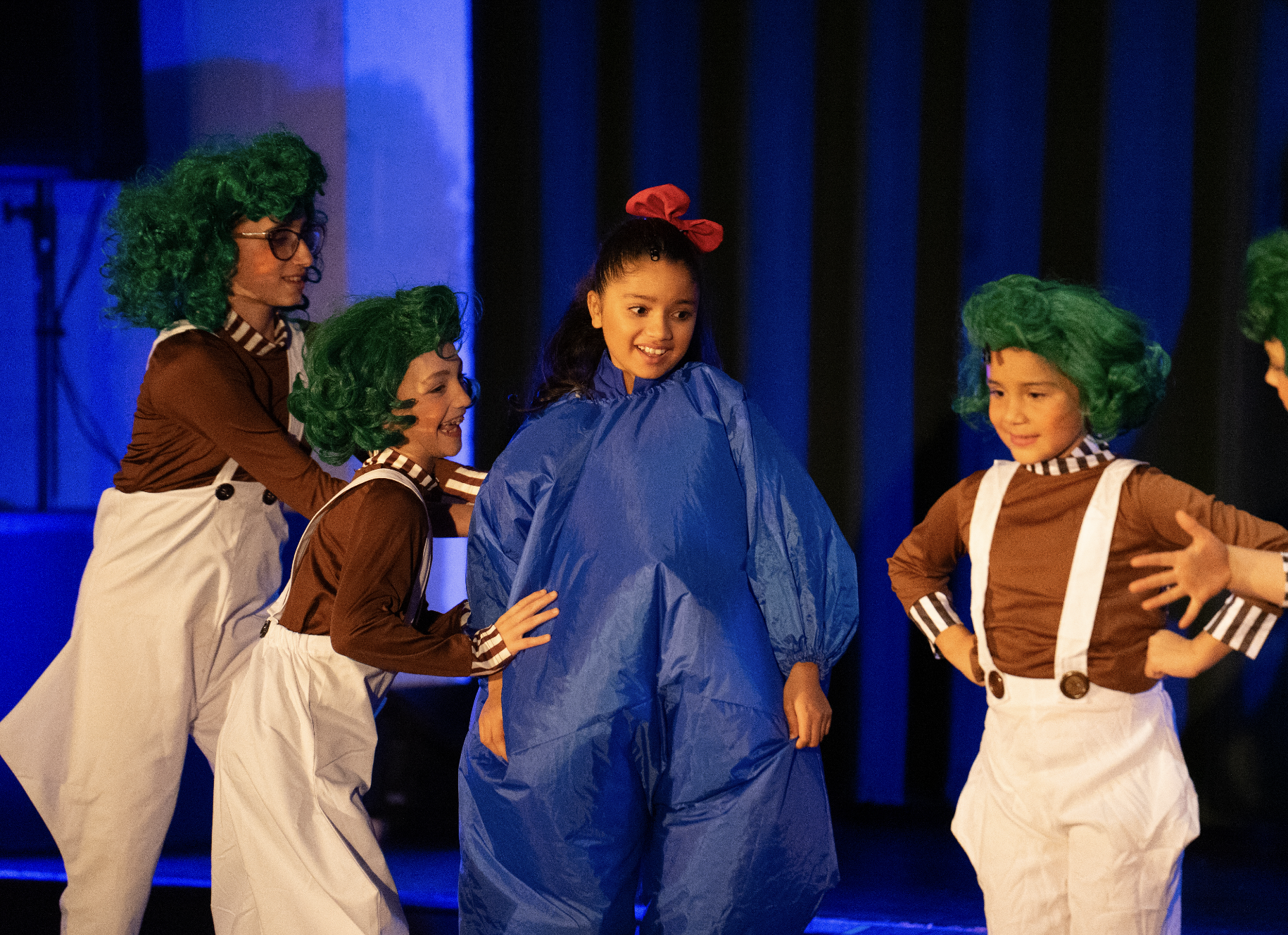 Children in costumes on stage, with four girls dressed as little beans with green wigs and suspenders and glasses, and a girl in a blue raincoat with a red bow in her hair, stage setup in background.