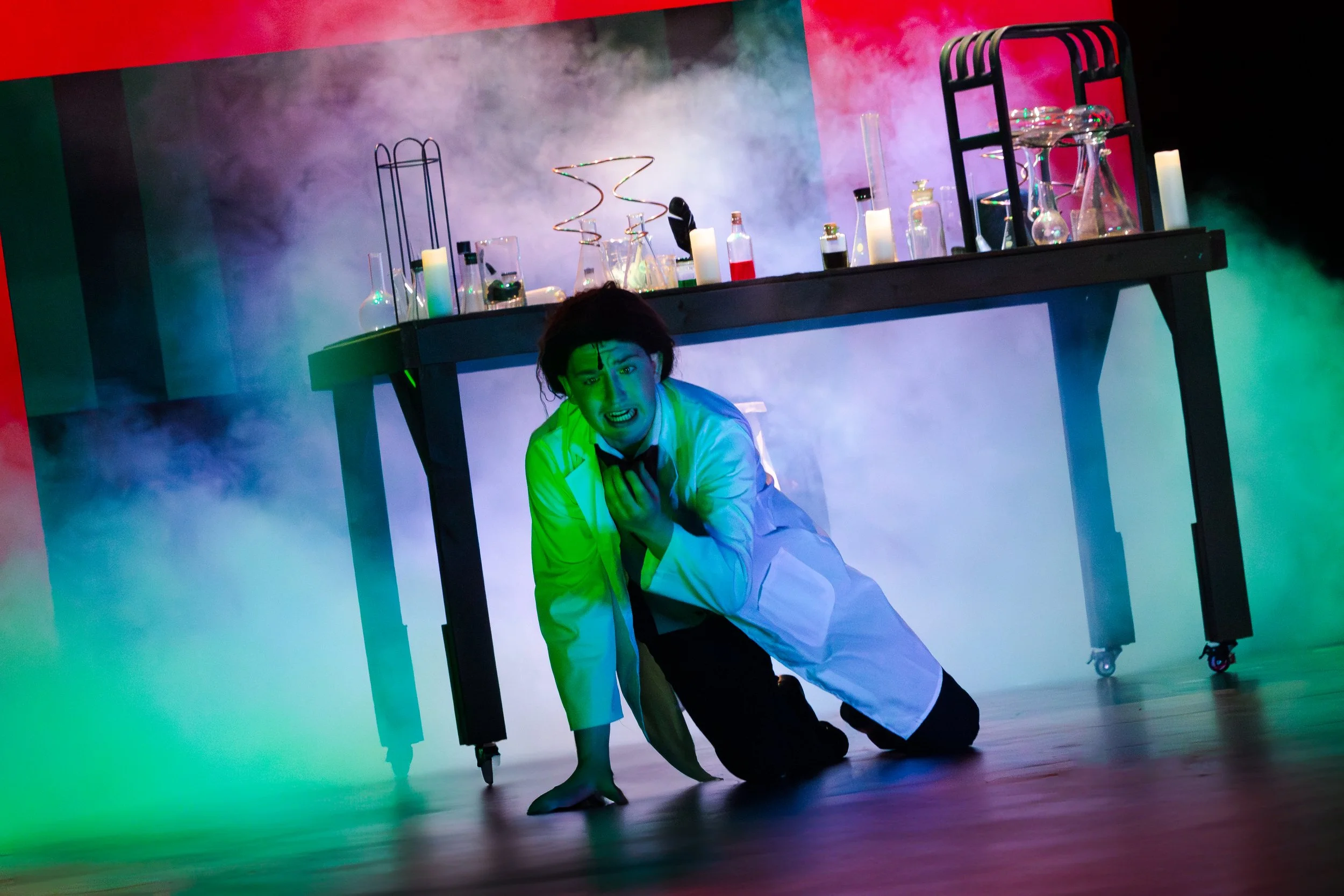 A person in a white lab coat kneels on the floor under a science table, gripping their chest with a distressed expression. The table is covered with laboratory glassware, and colorful smoke surrounds the scene, creating a dramatic, theatrical atmosph