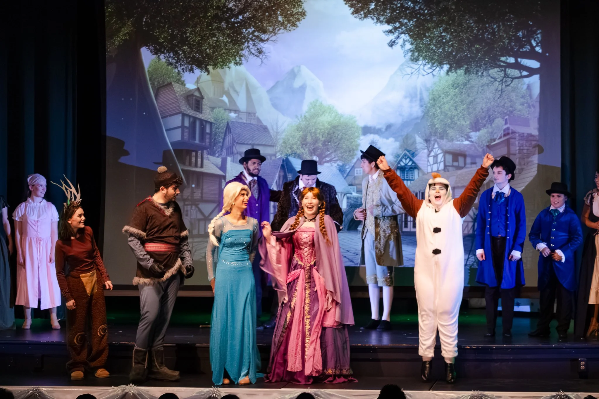 A theatrical stage performance with actors in colorful costumes, including a snowman costume with raised arms, and a backdrop of a scenic winter village with mountains.