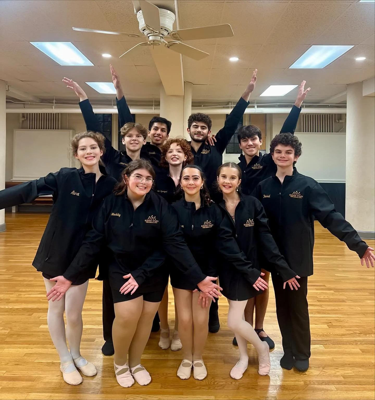 A group of ten young ballet dancers posing together in a dance studio, wearing black jackets with embroidered logos and their names, some in tights and ballet shoes, smiling and celebrating.