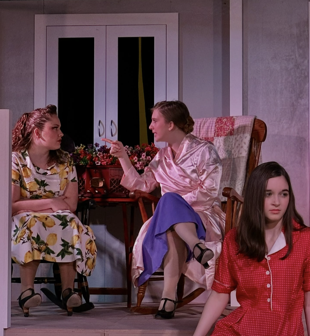 Three young women in vintage-style clothing engaged in a tense conversation in a living room. One is sitting on a rocking chair wearing a pink silk robe, another is sitting on a chair wearing a lemon print dress, and the third is sitting on the floor wearing a red checkered dress.