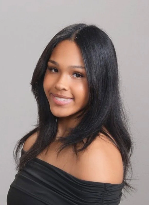A young man with long, curly hair, a mustache, and a light smile, wearing a striped shirt over a white t-shirt, posing against a neutral background.