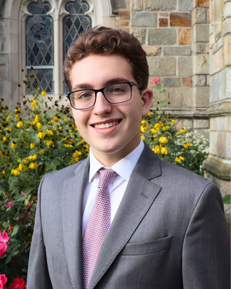 A young man with light skin, curly brown hair, and green eyes, smiling, wearing a pink button-up shirt and a dark blazer, sitting against a plain light-colored background.
