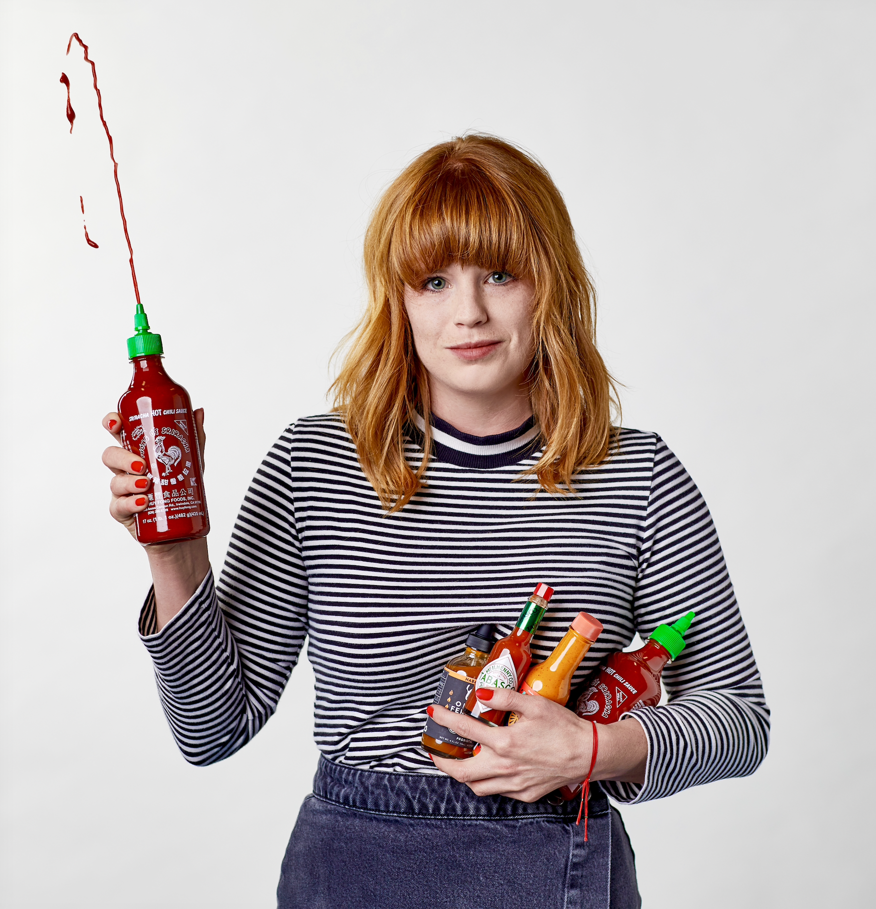 A woman with red hair and a striped shirt holds several bottles of condiments and sauce, including hot sauce, with one bottle in her hand with a splash hanging from the nozzle.