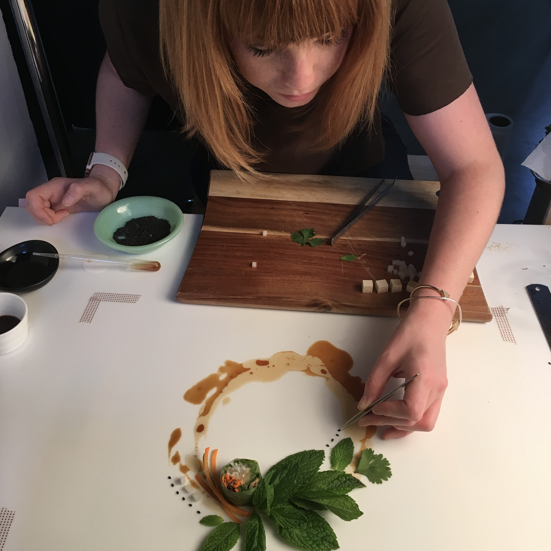 Woman with red hair creating an artful food presentation on a white surface, with green herbs, small vegetables, and sauce.
