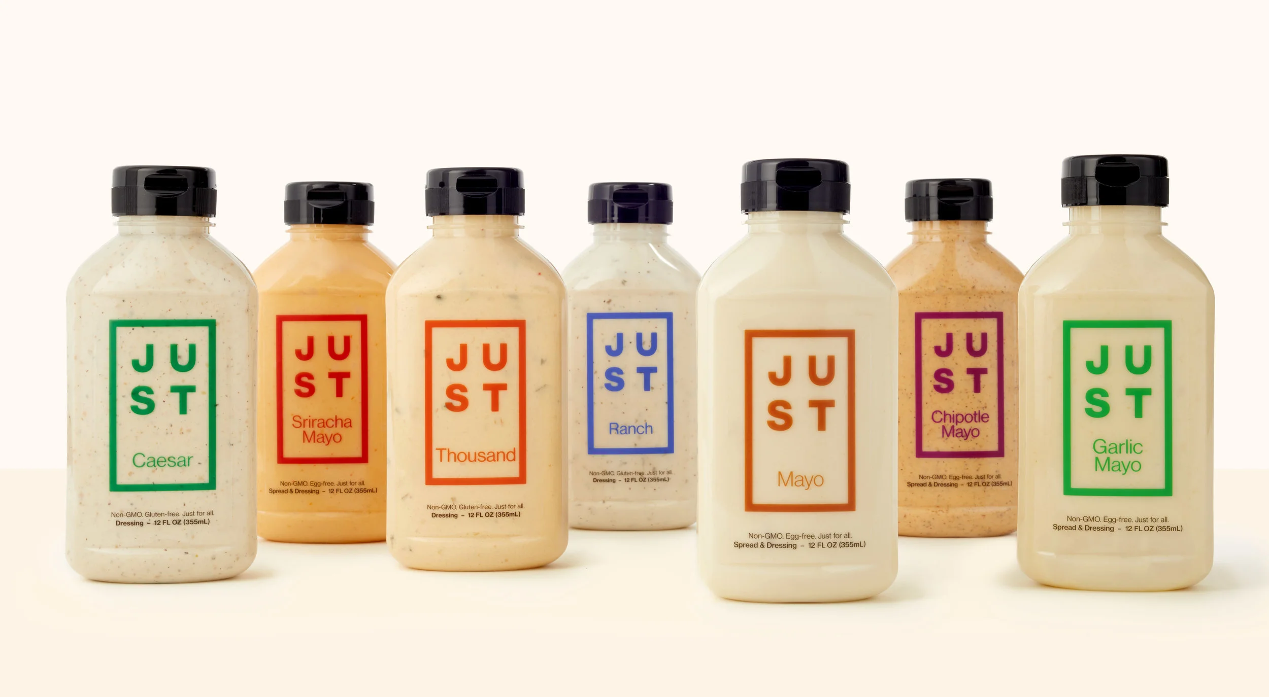 Six bottles of salad dressing with colorful labels reading 'JUST' and different flavors including Caesar, Sriracha Mayo, Thousand, Ranch, Mayo, and Garlic Mayo, arranged on a white surface.