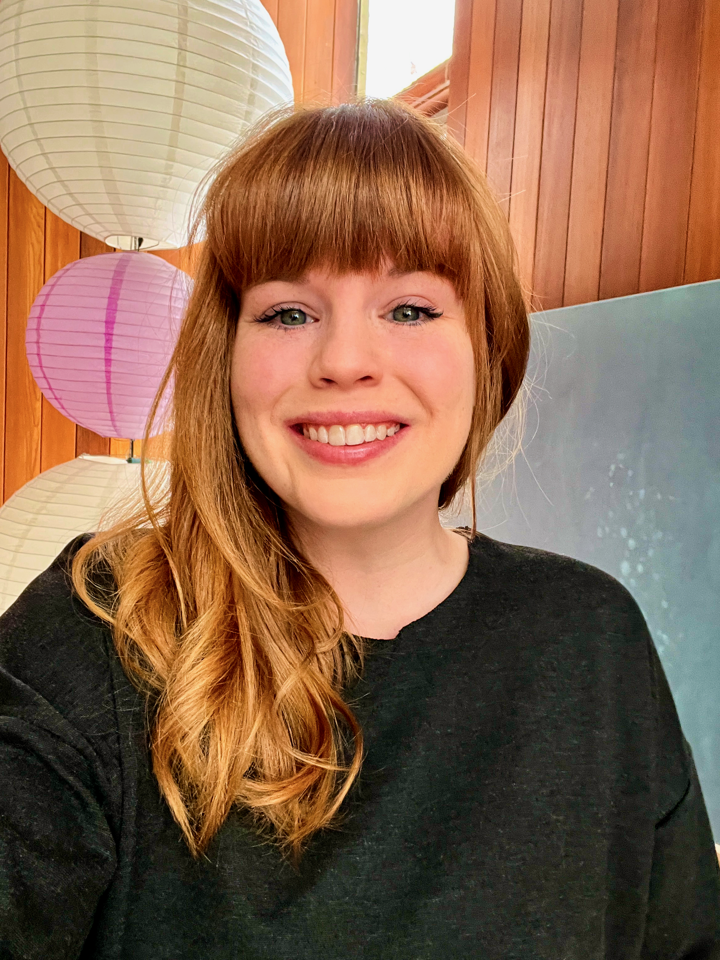 A young woman with long red hair and bangs, smiling, wearing a black top. Behind her are pink and white paper lanterns and a wooden wall.