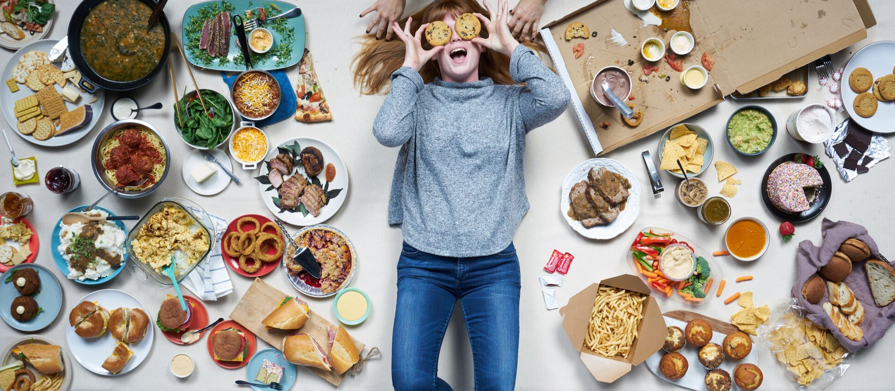 A woman lying on a table surrounded by various types of food, including pizza, pasta, salads, sandwiches, desserts, and snacks. She is holding cookies over her eyes and smiling.