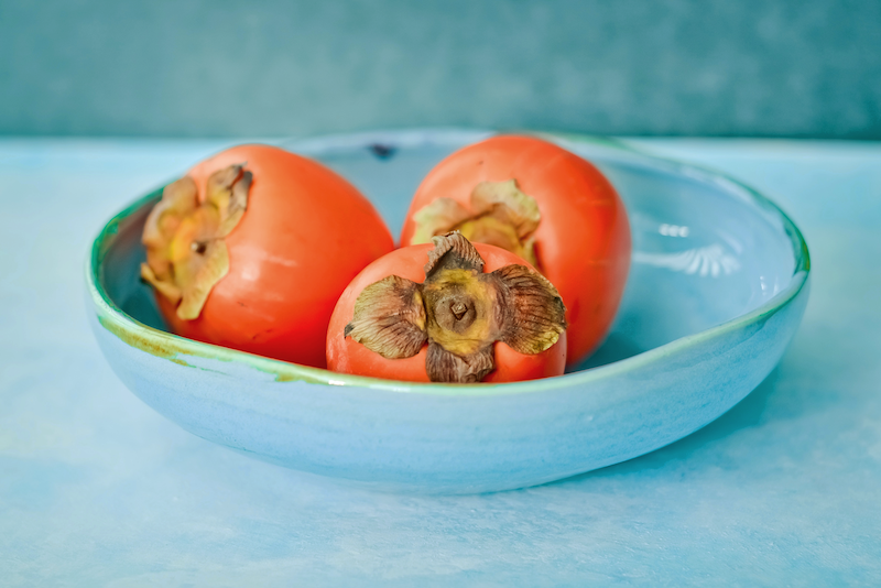 Three persimmon in a light blue bowl on a blue table