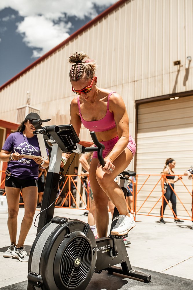 Woman in pink workout attire wearing sunglasses riding a stationary exercise bike outdoors, with other people and an industrial building in the background.