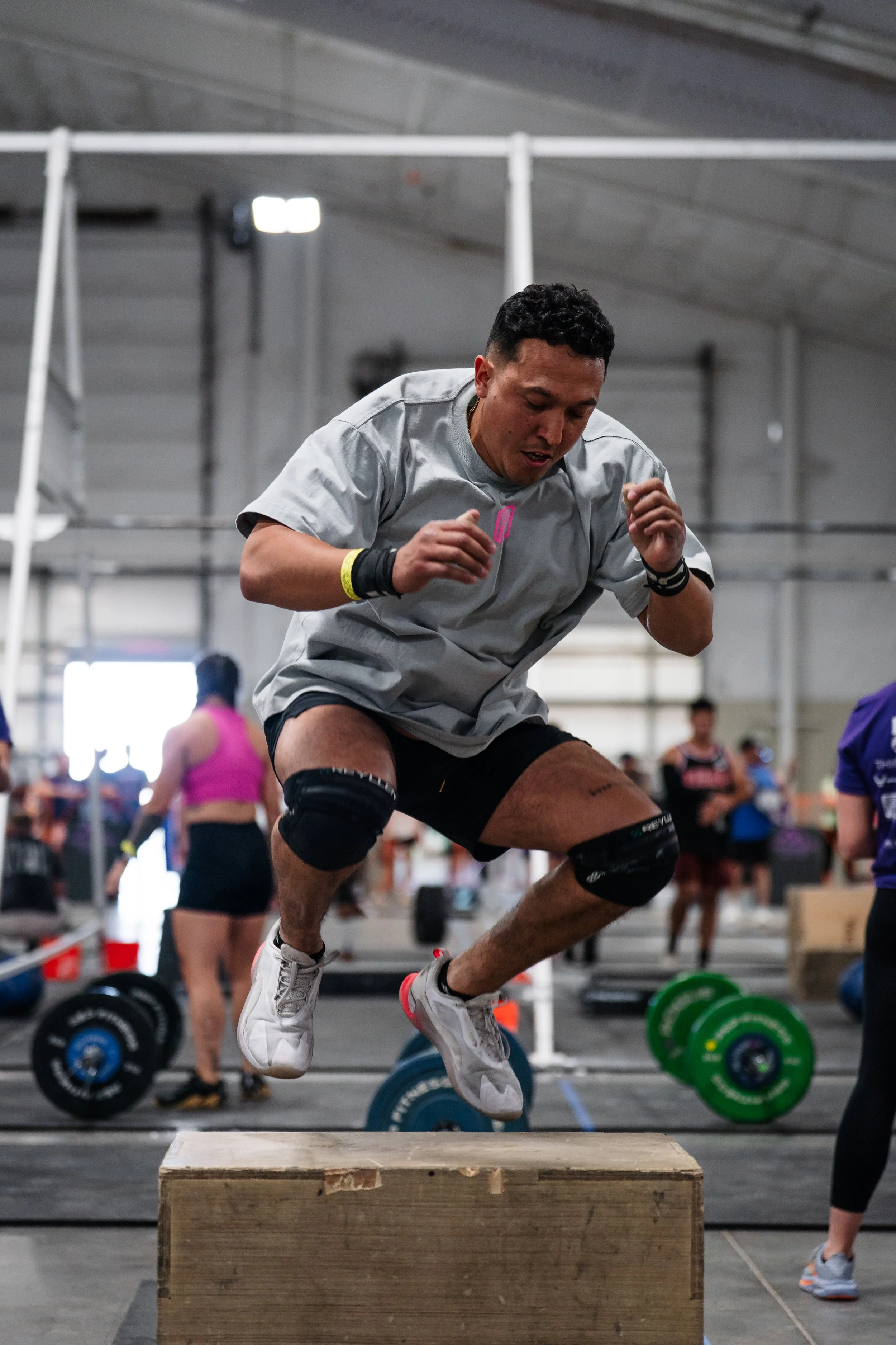 A shirtless man in gray shorts and sneakers lifting a dumbbell while balancing on a wooden platform during a workout session with a group of people standing behind orange barriers, watching him.