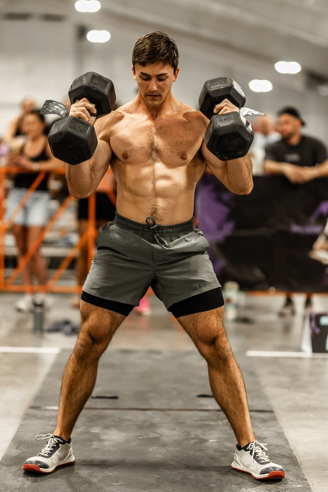 A muscular man lifting dumbbells in a gym, wearing shorts and sneakers, with a focused expression and a group of people in the background.