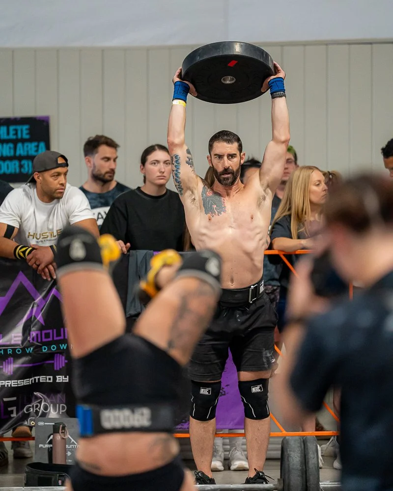 A muscular man is performing a back squat with a barbell loaded with weights at a gym or competition setting, surrounded by an audience of spectators.
