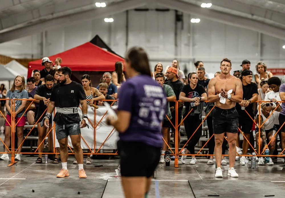 People gathered behind a barricade at a gym or indoor sports event, some with towels, water bottles, and athletic clothing, with a woman in purple in the foreground.
