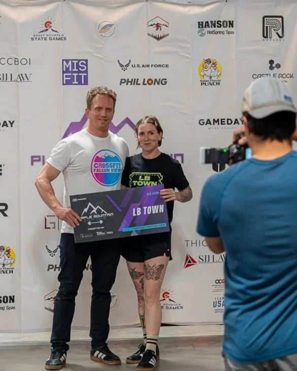 A man and a woman standing together, holding a large sponsor check at a CrossFit event, with a photographer taking their picture. The woman has tattoos and is wearing a black shirt, while the man is wearing a white T-shirt. The background displays multiple sponsor logos and event branding.
