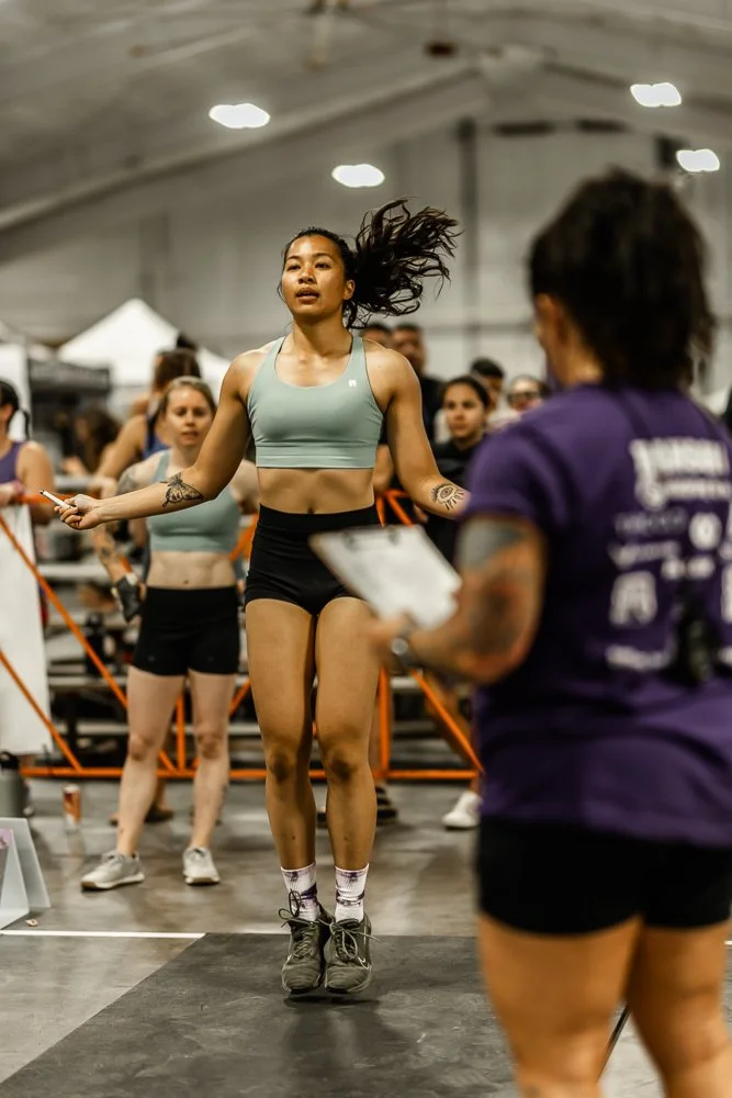A woman in a pink sports top and black shorts smiling after lifting weights in a gym with other people in the background.
